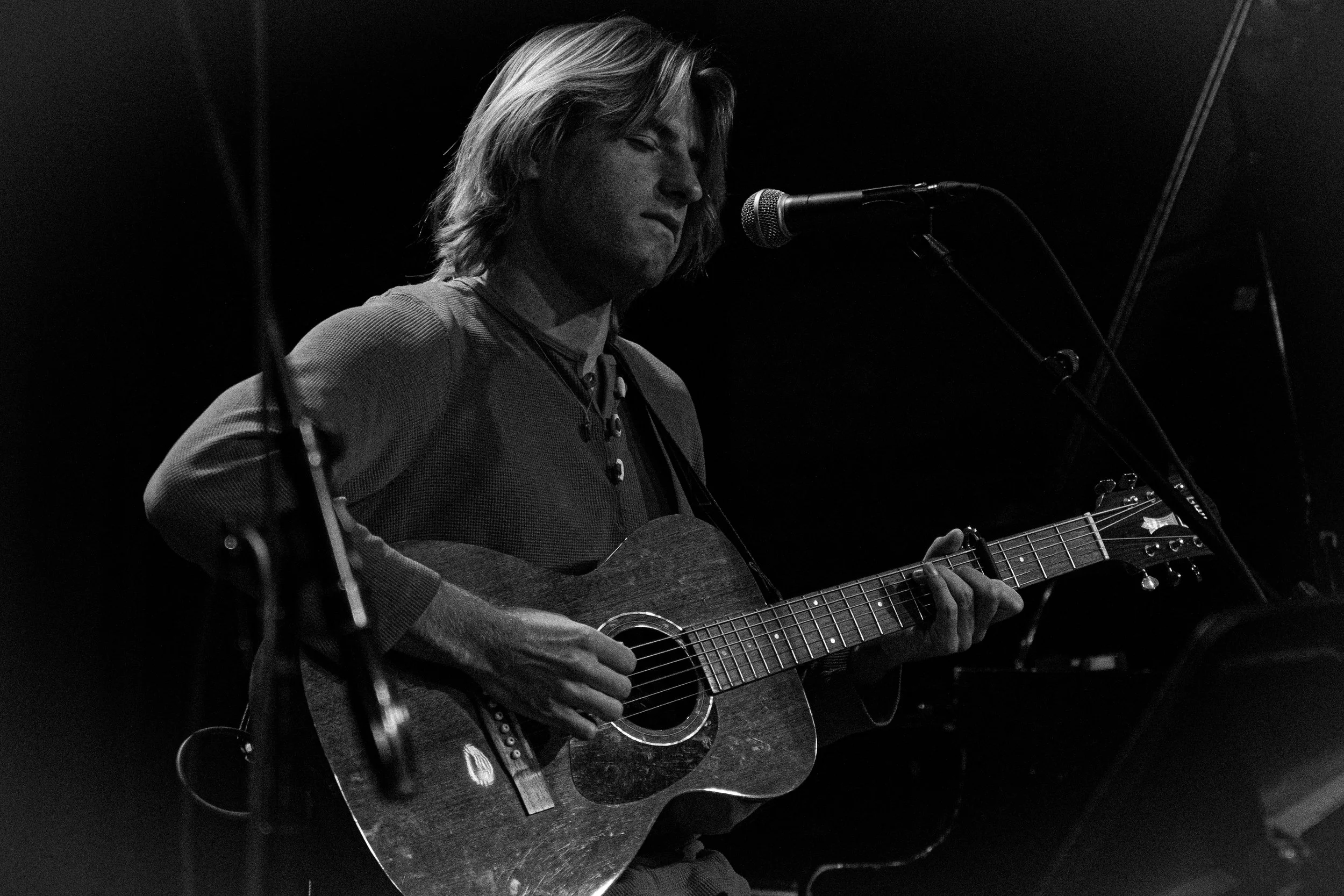 A black and white photo of Jack Corliss playing an acoustic guitar and singing into a microphone on a dark stage at SoHo Music House in Santa Barbara.