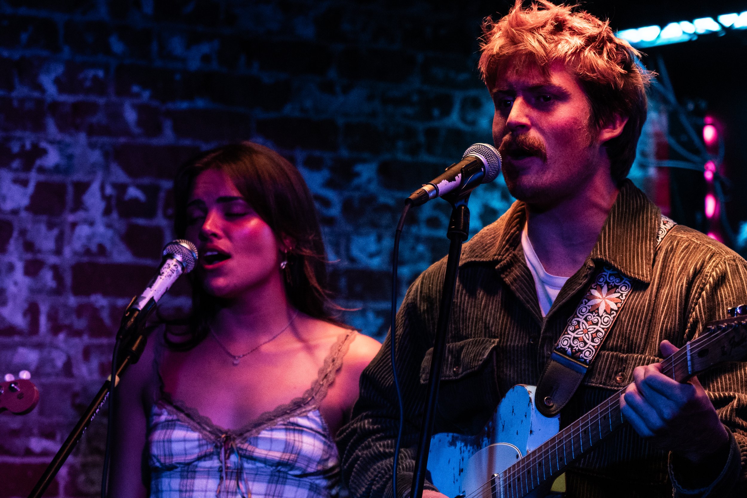Dolly Vaarden singing and playing guitar on stage with brick wall background, colorful stage lighting, and microphones at Whiskey Richards Santa Barbara.