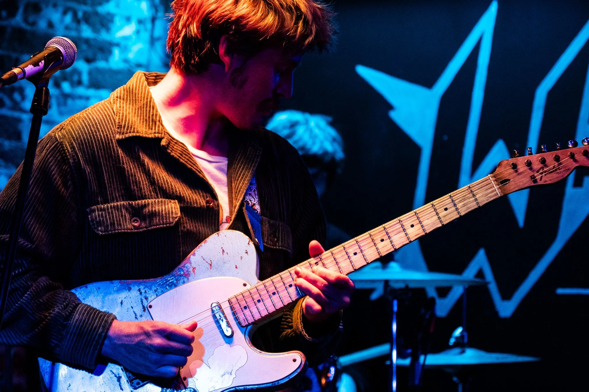 A young man in a dark jacket playing an electric guitar on stage with blue and purple lighting, with a microphone to his left and a brick wall in the background.