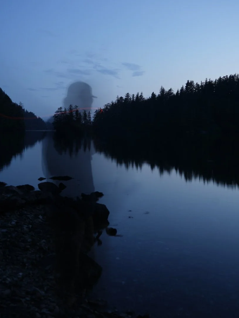 A lake at dusk surrounded by trees, with a faint reflection of the sky on the water. A person’s silhouette is visible in the sky, blending with the clouds.