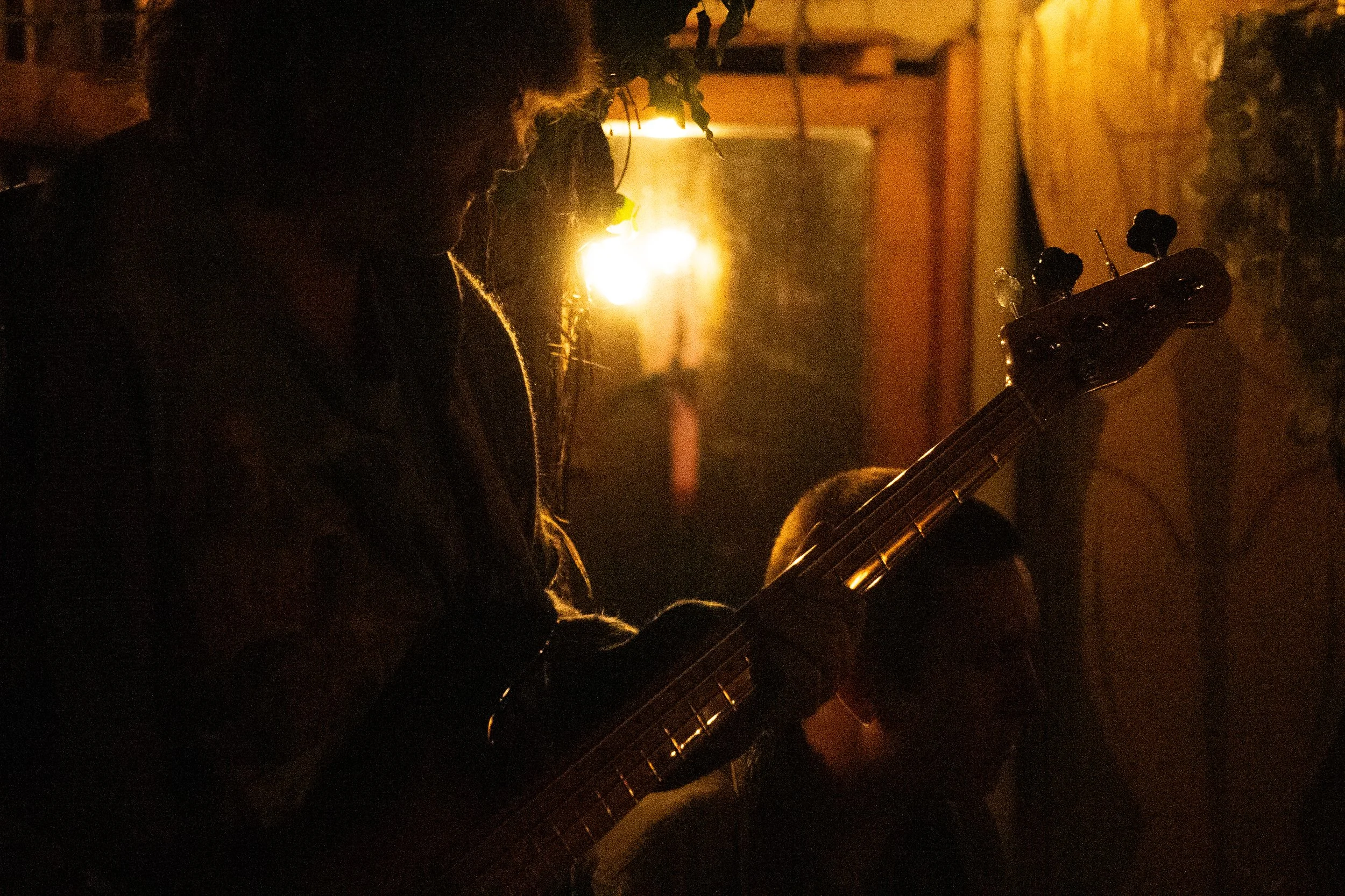 Silhouette of a person playing guitar in dim, warm lighting, with another person visible in the shadowed background.