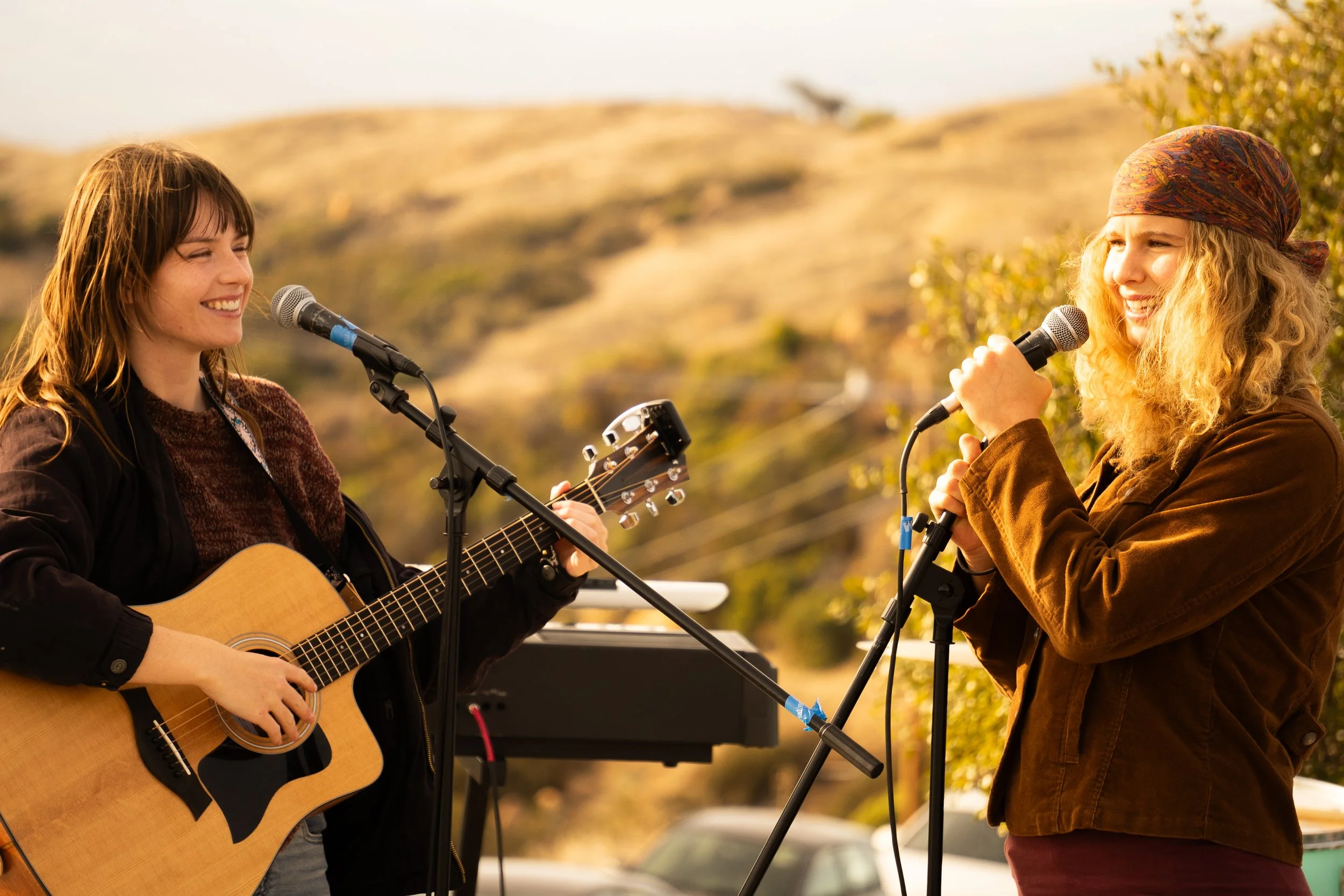 Two women performing music outdoors with microphones; one is playing acoustic guitar, and the other is singing.