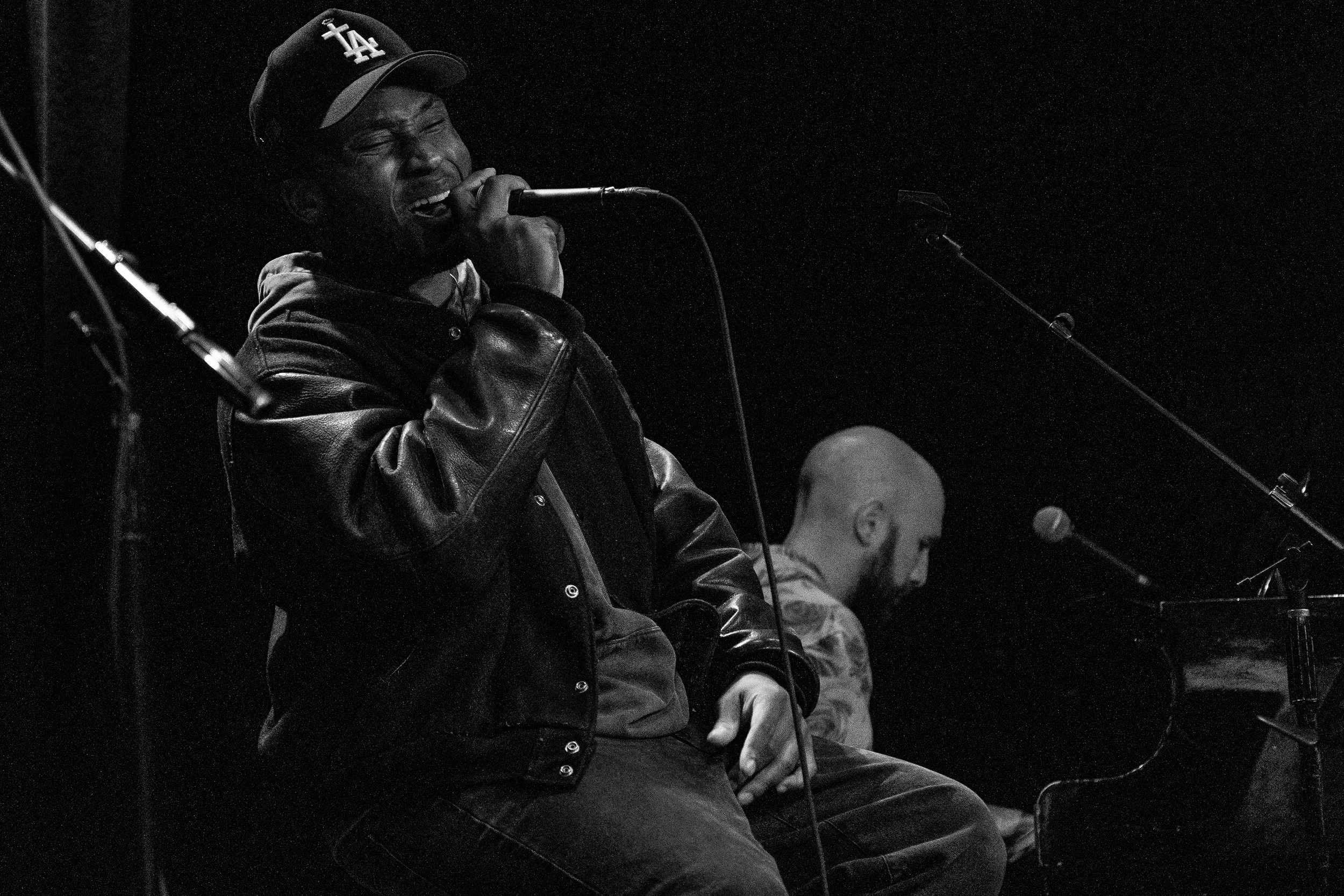 Singer Brayelle wearing a baseball cap and leather jacket performing into a microphone, with a pianist playing piano in the background, at Soho House Santa Barbara