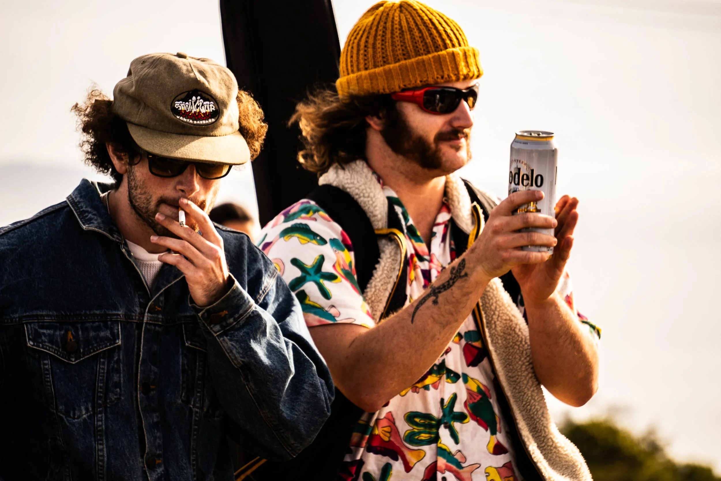 Two men outdoors, one smoking a cigarette, the other holding a can of Modelo beer, wearing a multicolored Hawaiian shirt, a yellow knit beanie, and a backpack, both of them watching a mountain show above Santa Barbara.