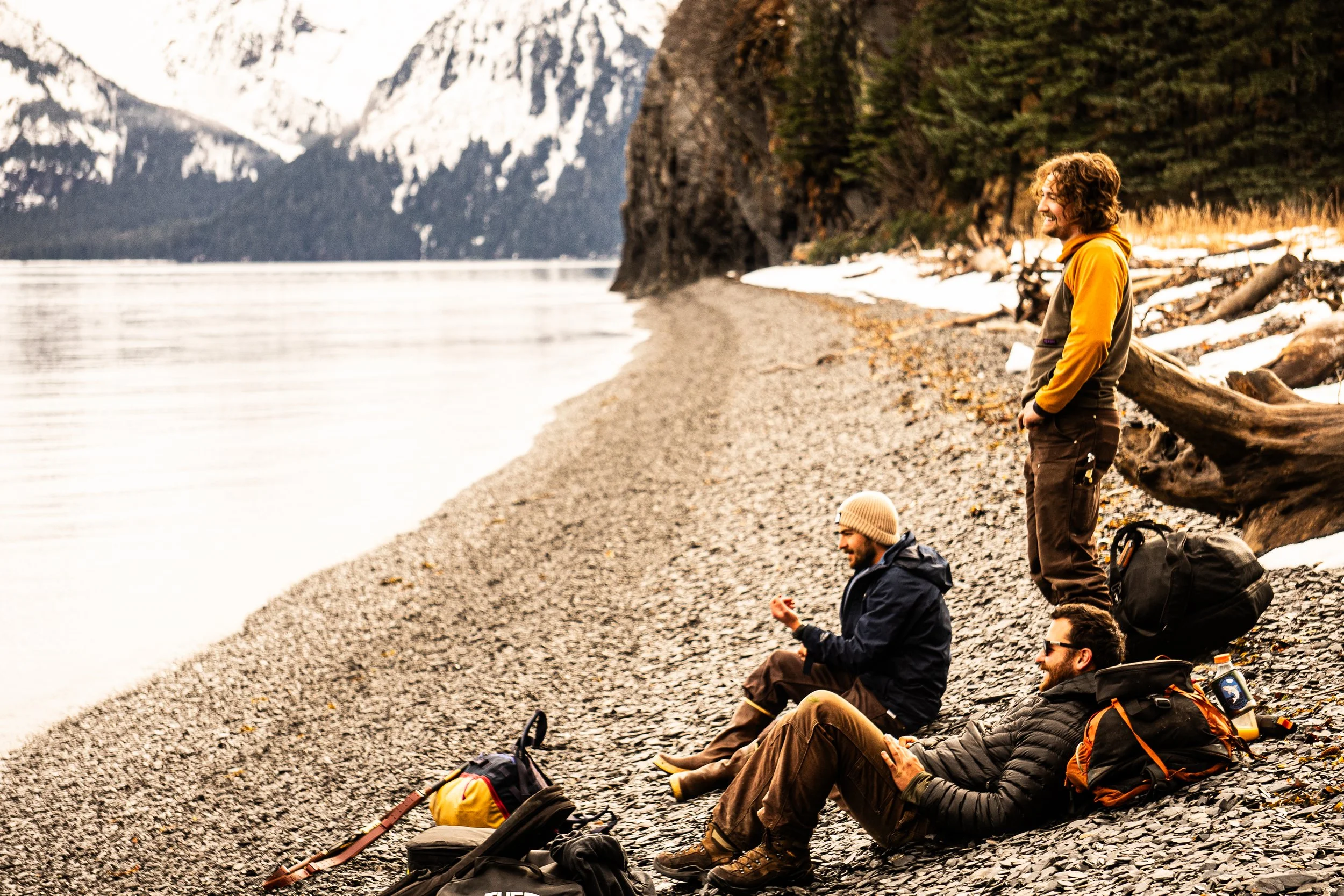 Three friends relaxing on a rocky beach near a body of water, with snowy mountains and pine trees in the background. They are sitting and standing with backpacks and outdoor gear.