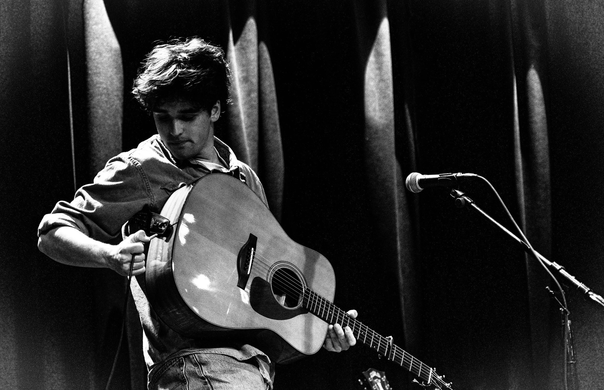 Miles Carter is holding an acoustic guitar on a stage, looking down at the guitar at SoHo Santa Barbara. There is a microphone on a stand in front of him, and dark curtains in the background.