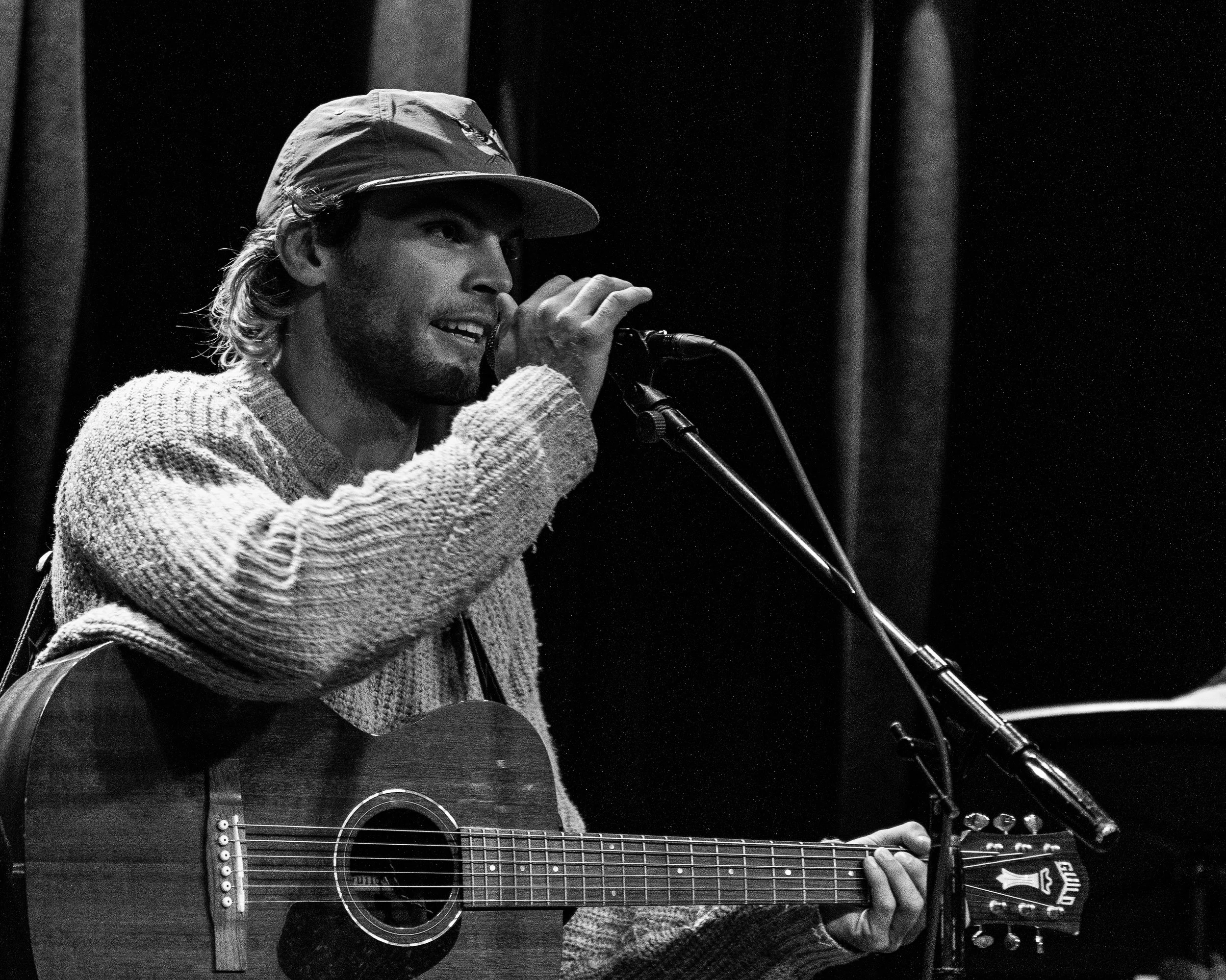 Moe Johnson performing on stage with a guitar, singing into a microphone, wearing a cap and a sweater.