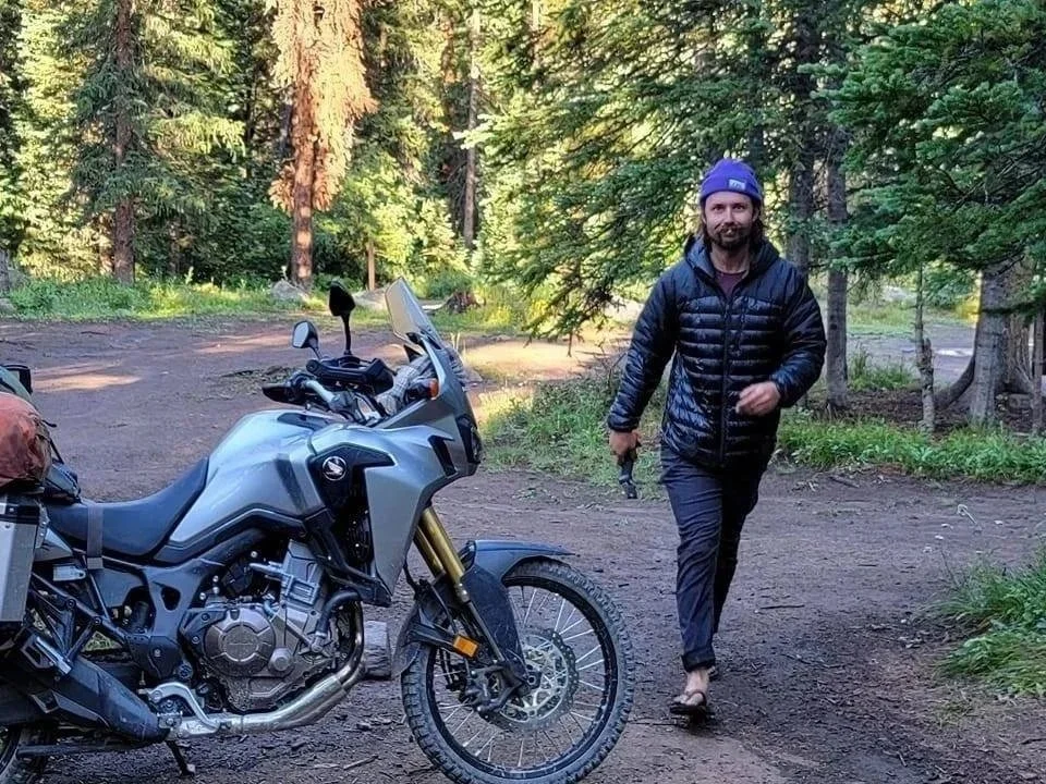 A man in a blue beanie and black jacket walking on a dirt trail next to a silver adventure motorcycle in a forest setting.