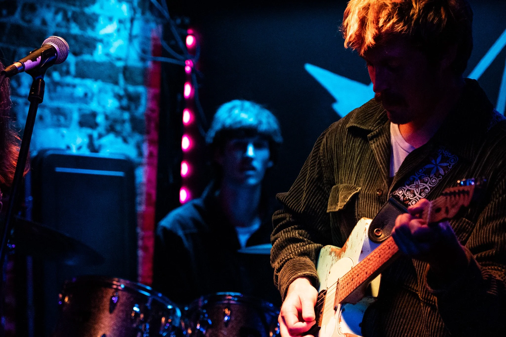 Someone playing the guitar on stage with a drummer in the background, dimly lit with colorful stage lighting.