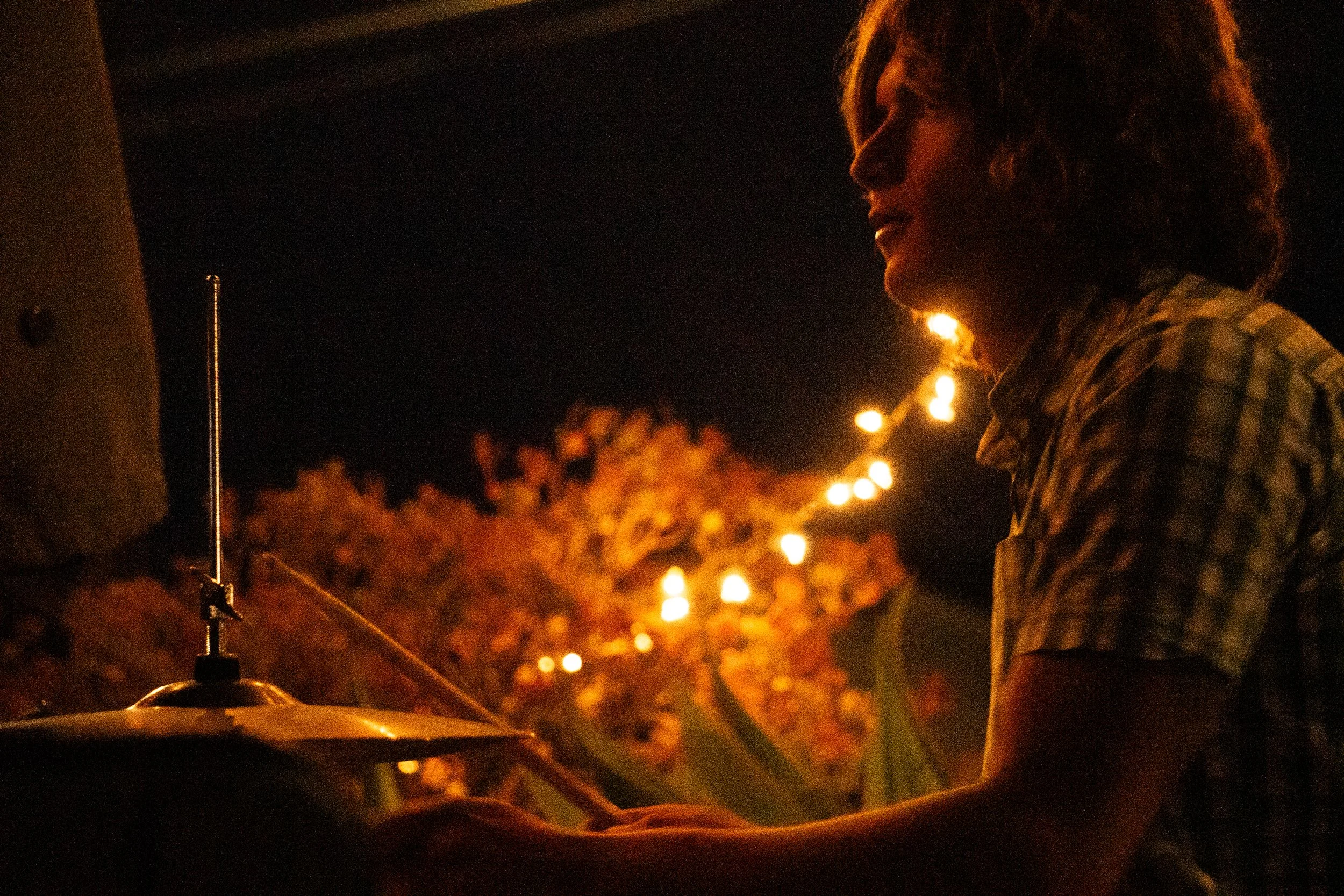 A person playing the drums at night, with string lights in the background.