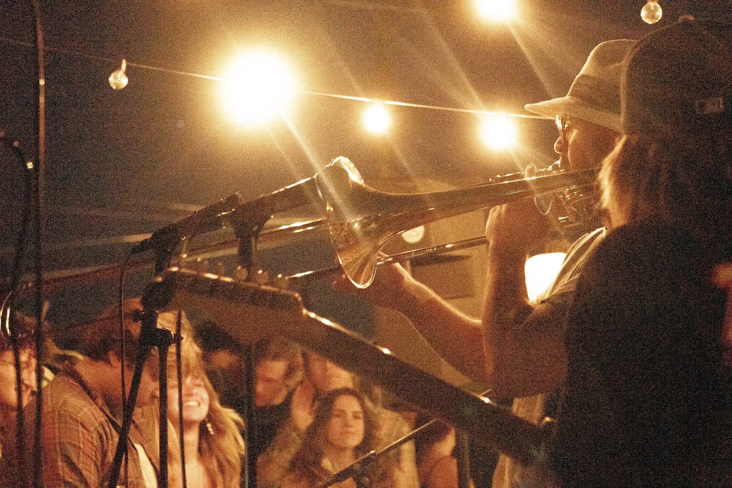 A person playing a trumpet at night under string lights, with a crowd of cheerful people watching.