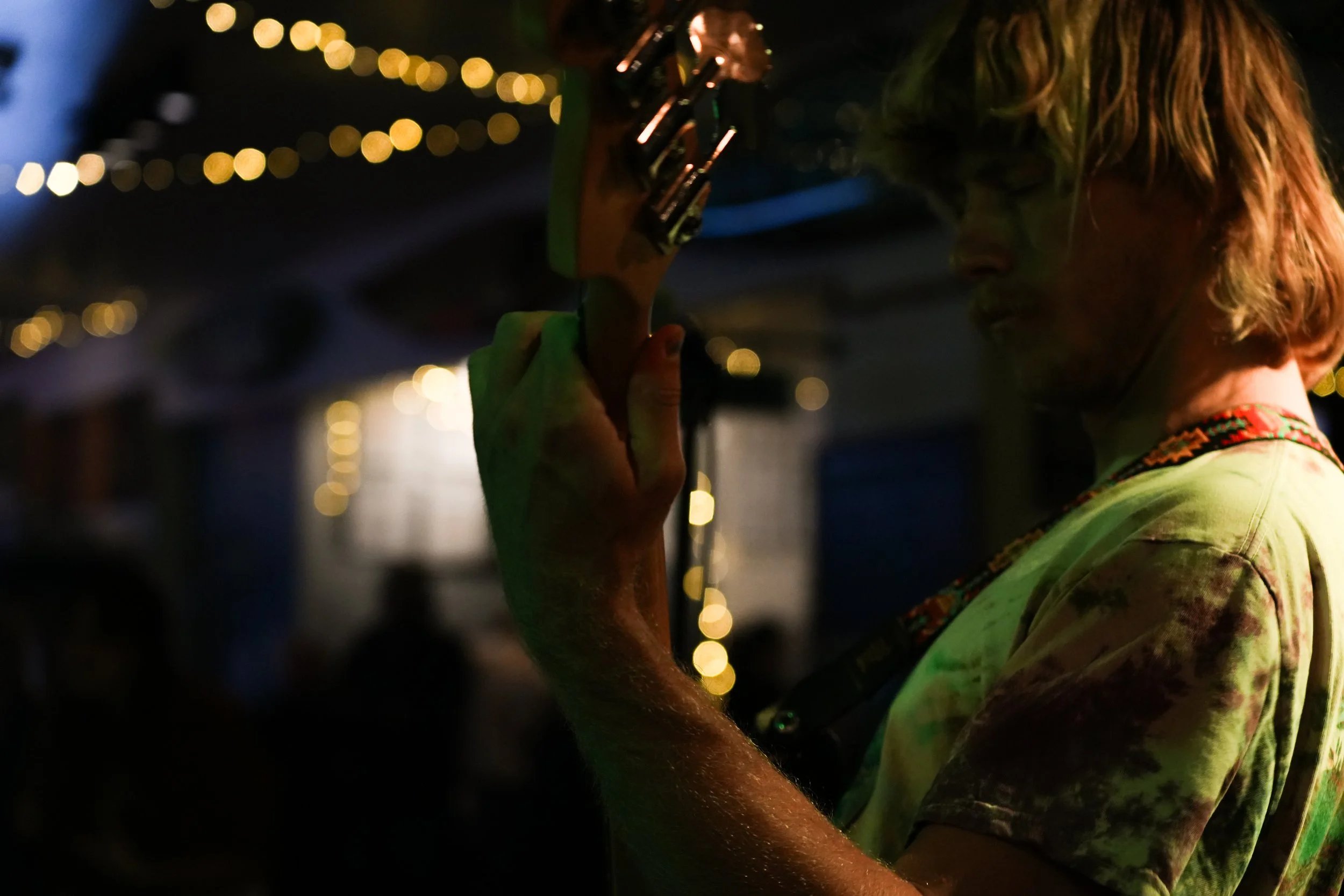 A person with shoulder-length hair playing an electric guitar in a dimly lit setting with string lights in the background.