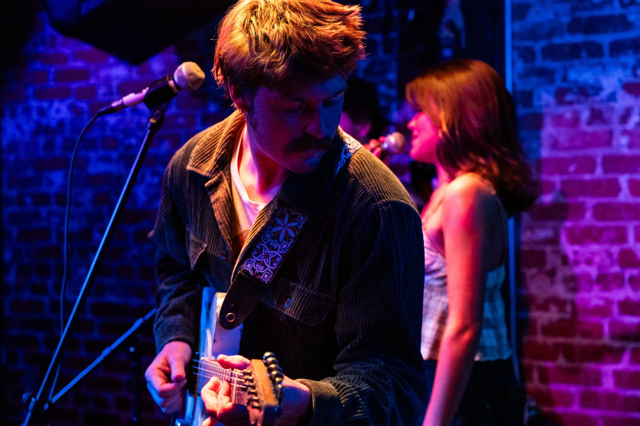 A male musician with a mustache playing an electric guitar on stage, with a female singer in the background, against a brick wall illuminated with colorful stage lights.