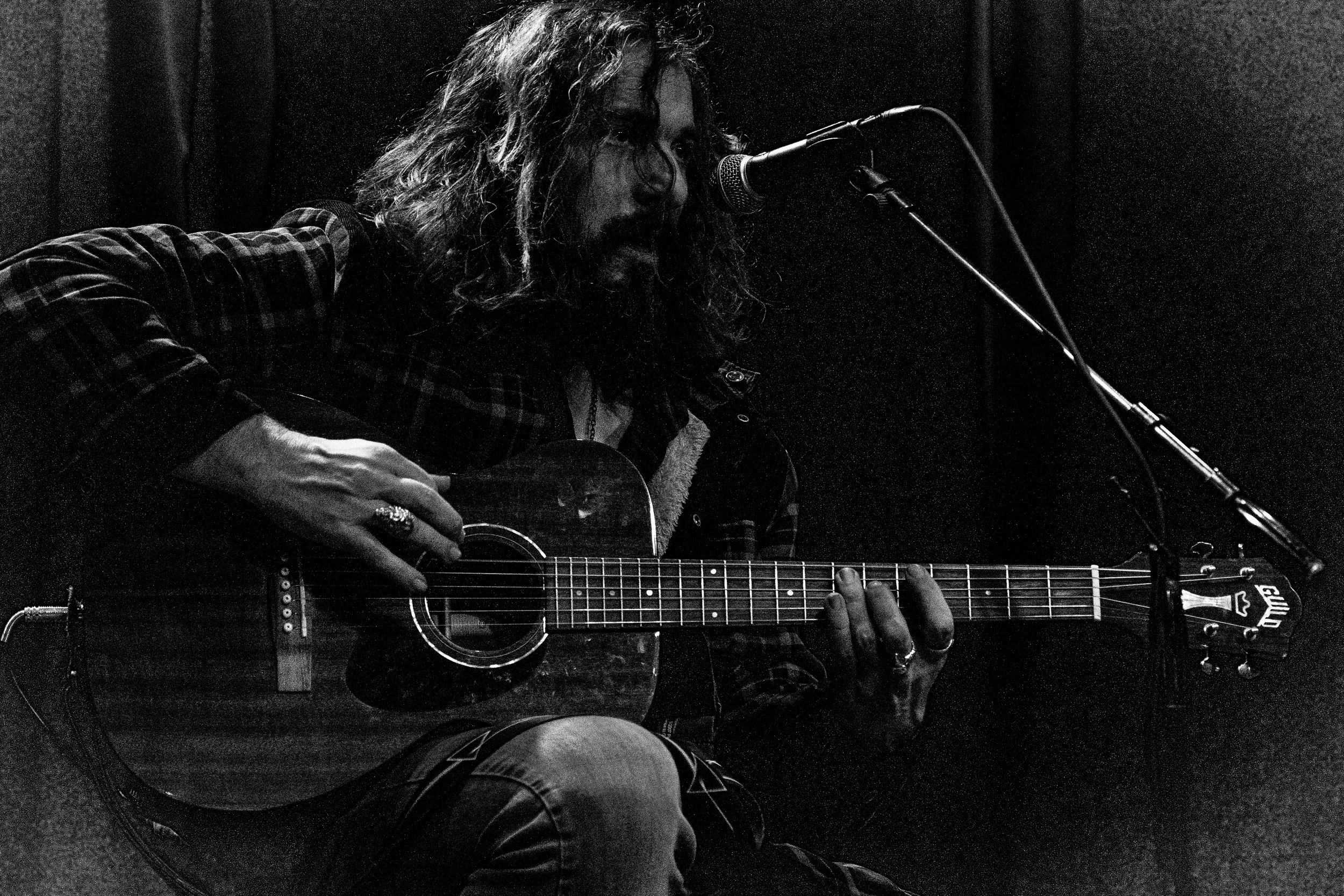 Black and white photograph of Wesley Birch with long hair and beard playing an acoustic guitar, sitting in front of a microphone with a dark background.