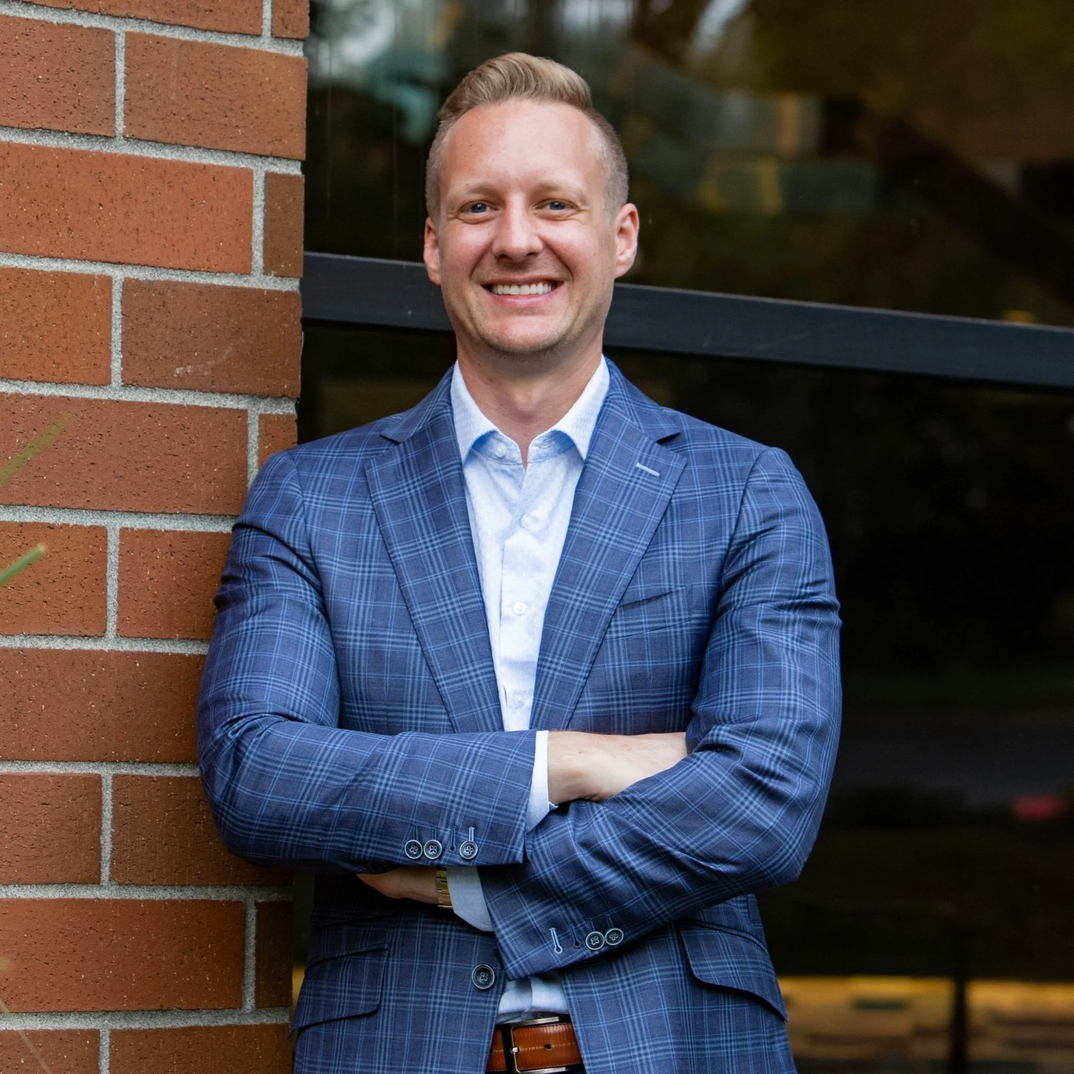 A man in a blue plaid suit jacket and light blue dress shirt standing outdoors, leaning against a brick wall, smiling with arms crossed.