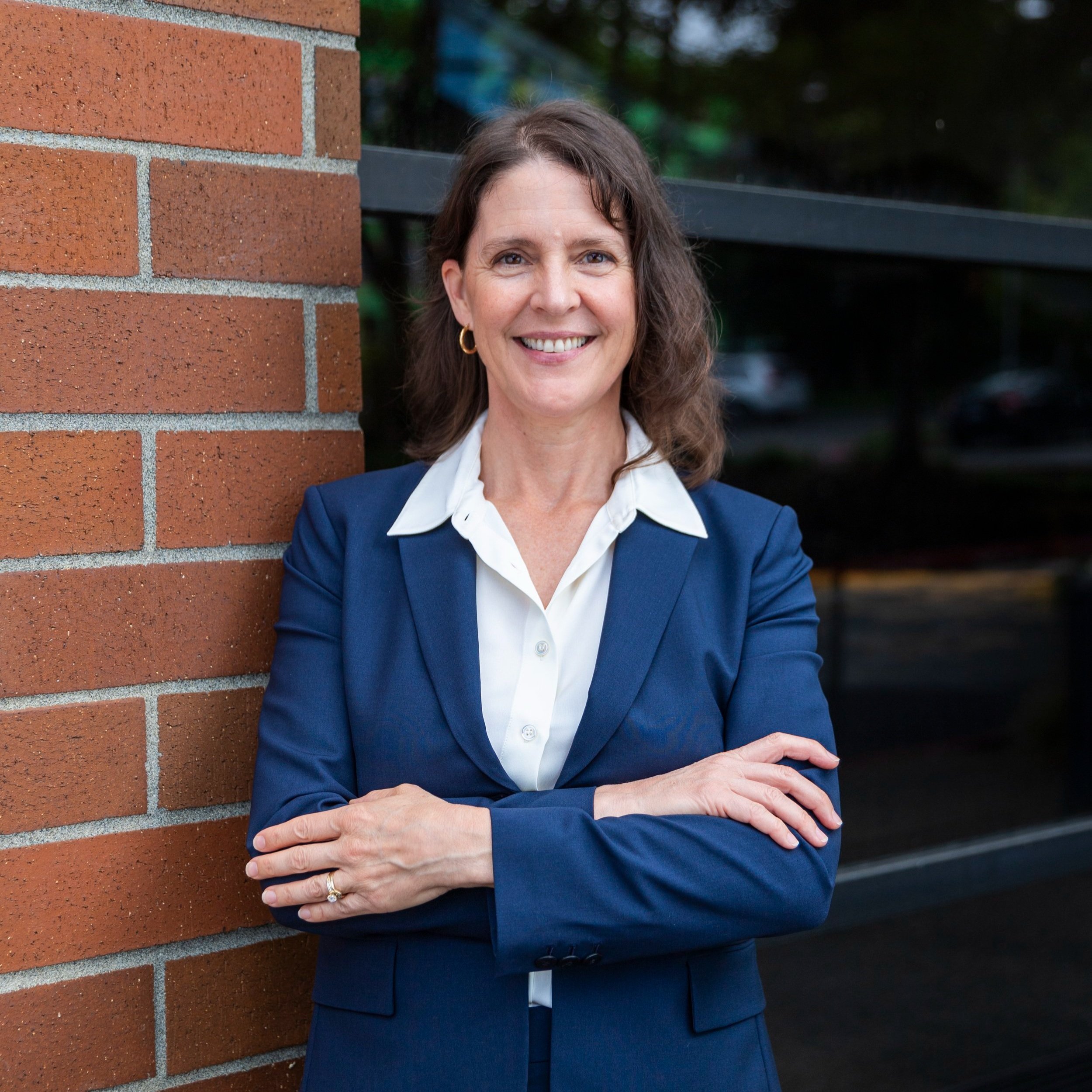 A woman in a navy blazer and white shirt standing outdoors against a brick wall, smiling with arms crossed.
