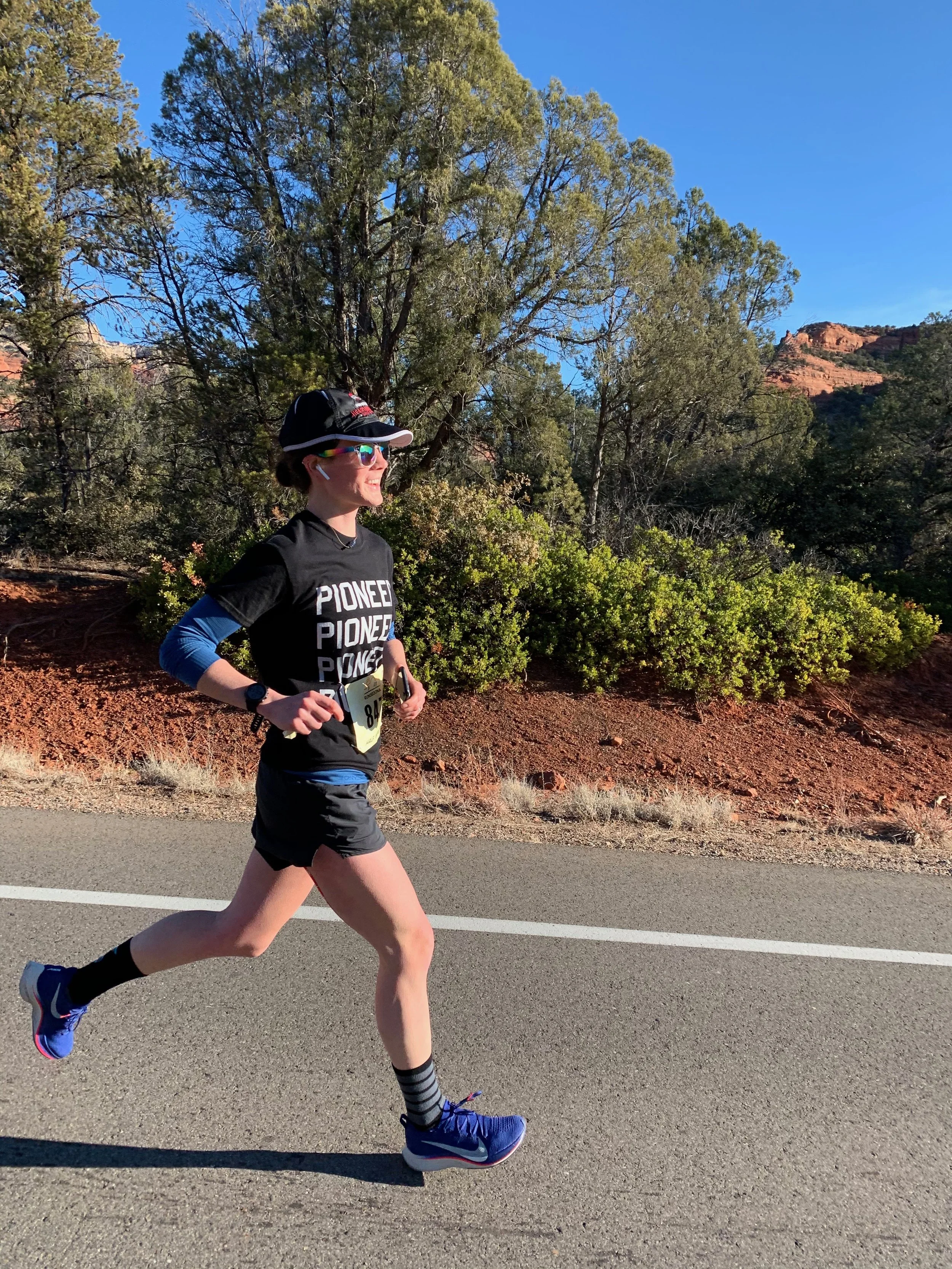 A woman running in a race on a paved road with trees and red rocks in the background.