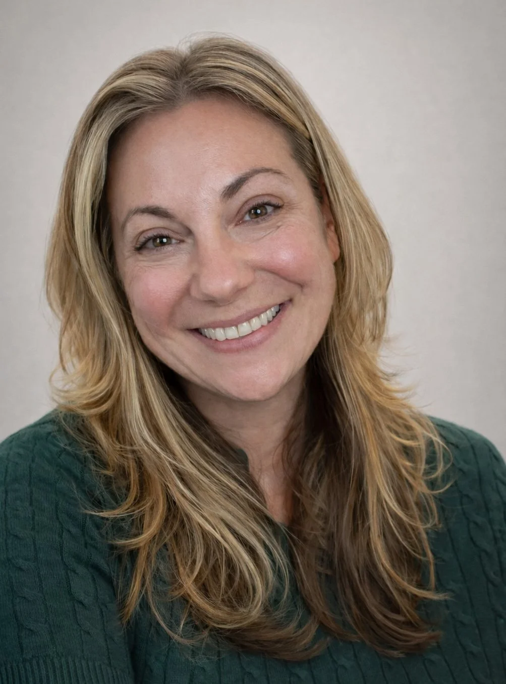 A woman with long, wavy blonde hair smiling at the camera, wearing a white turtleneck against a light peach background.