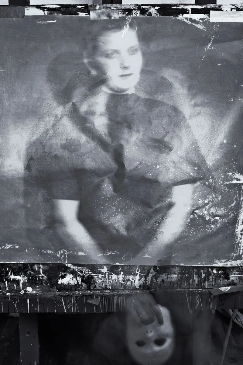 A black and white painting of a young woman with a blurred face, sitting with her hands on her lap, with an abstract background. The painting is reflected on a shiny surface below.
