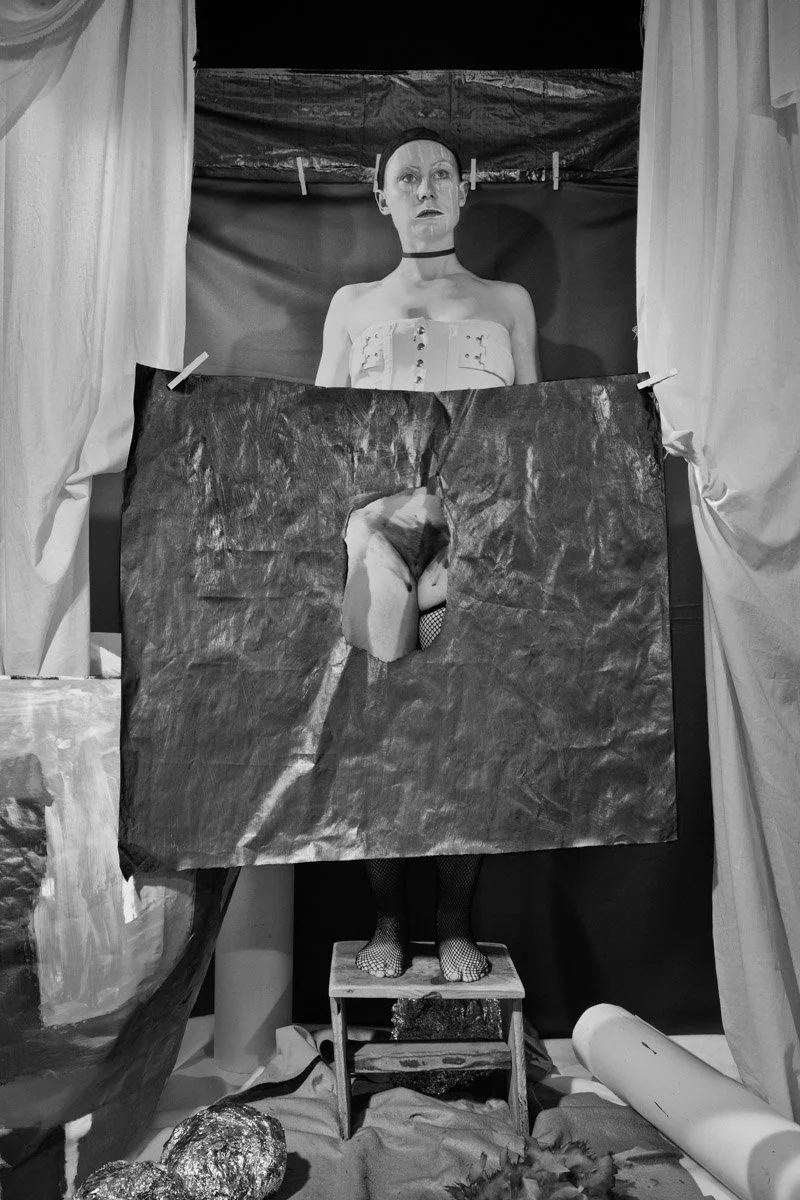 A black and white art installation features a mannequin or sculpture of a woman with a choker, wearing a strapless top, behind a torn paper sheet. The paper has a cutout revealing her legs, which are covered in fishnet stockings, and her feet are on