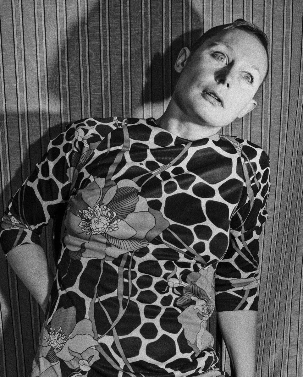 A woman with short hair and light skin sitting against a wooden panel wall. She is wearing a patterned shirt with large floral and animal print designs. Her head is tilted slightly, and she gazes upward with an open-mouthed expression.