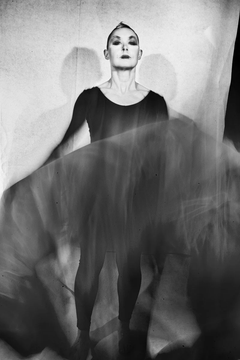 Black and white photo of a woman with styled hair and makeup, wearing a dark top, standing in front of a plain background, with a large leaf or fabric partly covering her lower body.