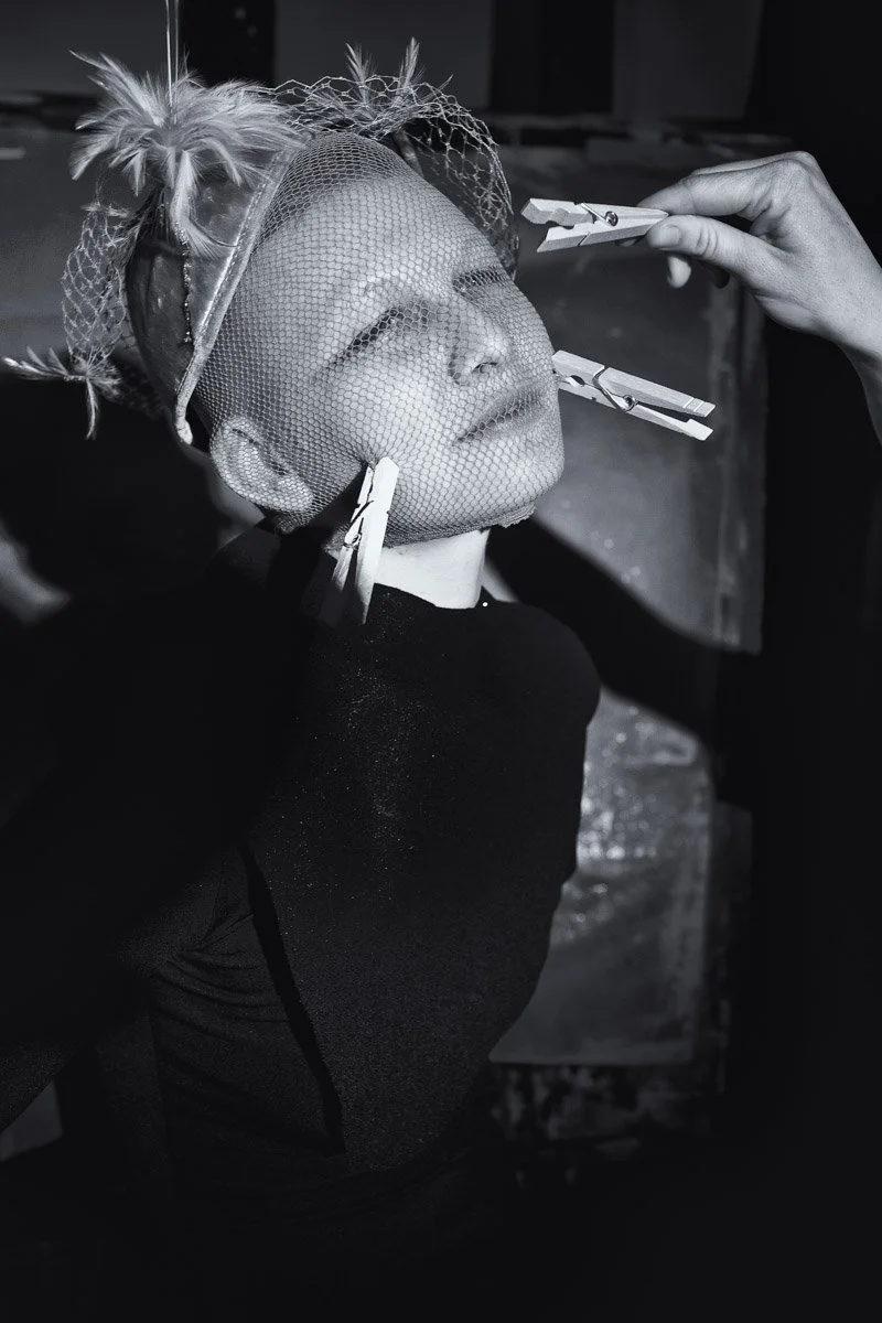 A person with makeup or prosthetics is having the lower part of their face painted or adjusted while their face is covered with a fine mesh net and they wear a headpiece with feathers, during a creative or theatrical makeup process.
