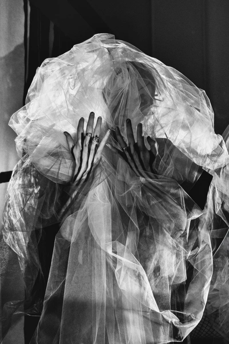 A person enveloped in layered, sheer fabric creating a cloud-like effect, with hands touching the fabric and partially obscured face visible through the material.