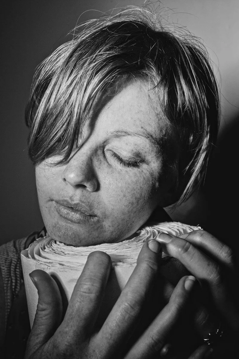 A woman with short hair rubbing her face and eyes closed, holding a large rolled object close to her face in black and white.