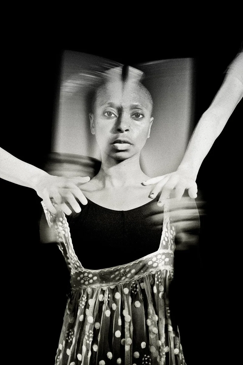 A black and white photo of a woman with a blurred mirror reflection above her face, and hands adjusting her dress from behind, creating a surreal artistic effect.