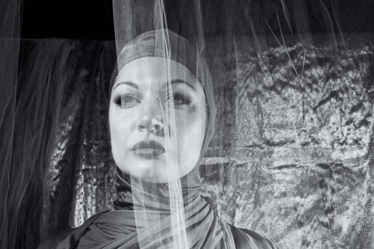A woman with makeup and a headscarf looking through a sheer fabric with a stone wall in the background.