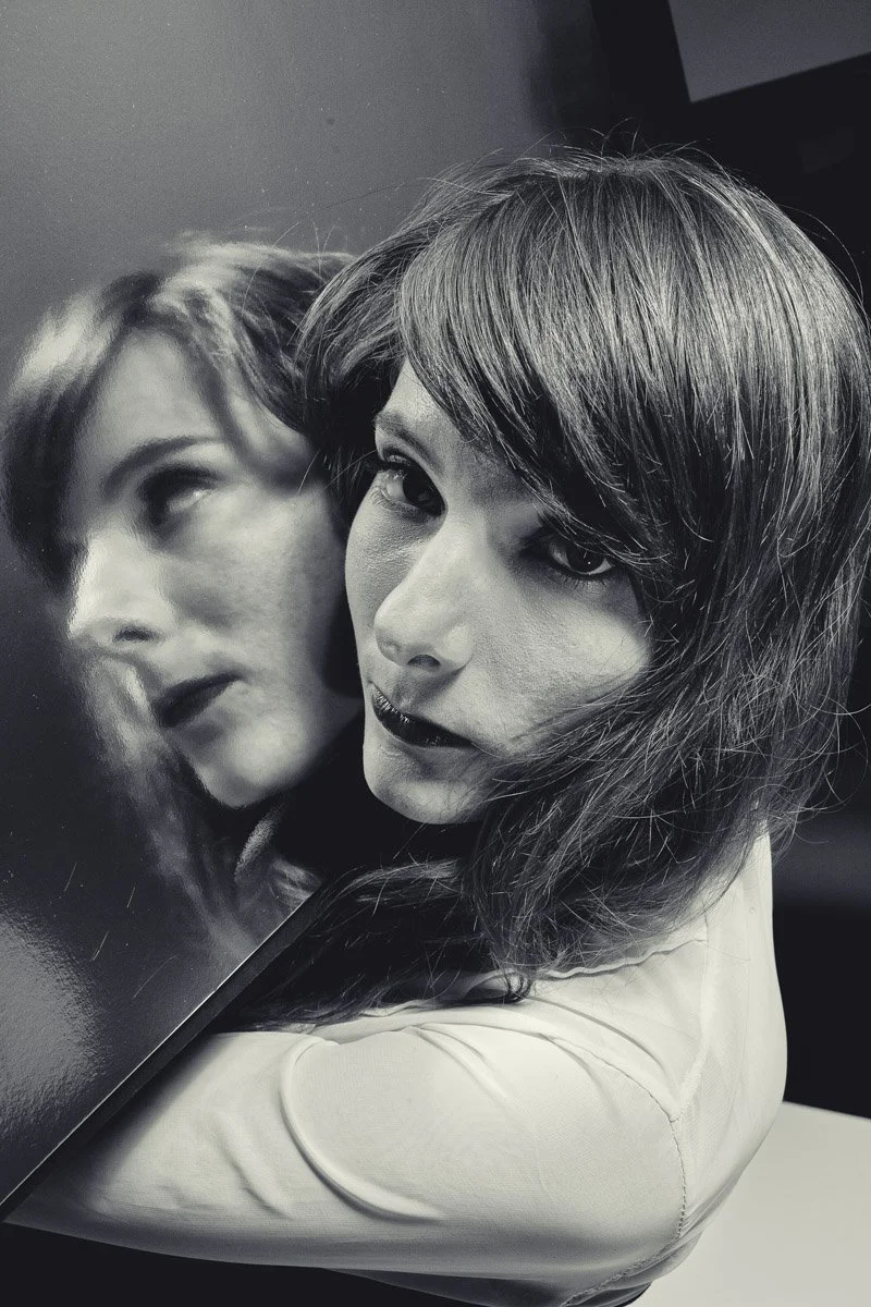 Black and white photo of a woman holding a mirror that reflects her face, showing her with dark lipstick and styled hair.