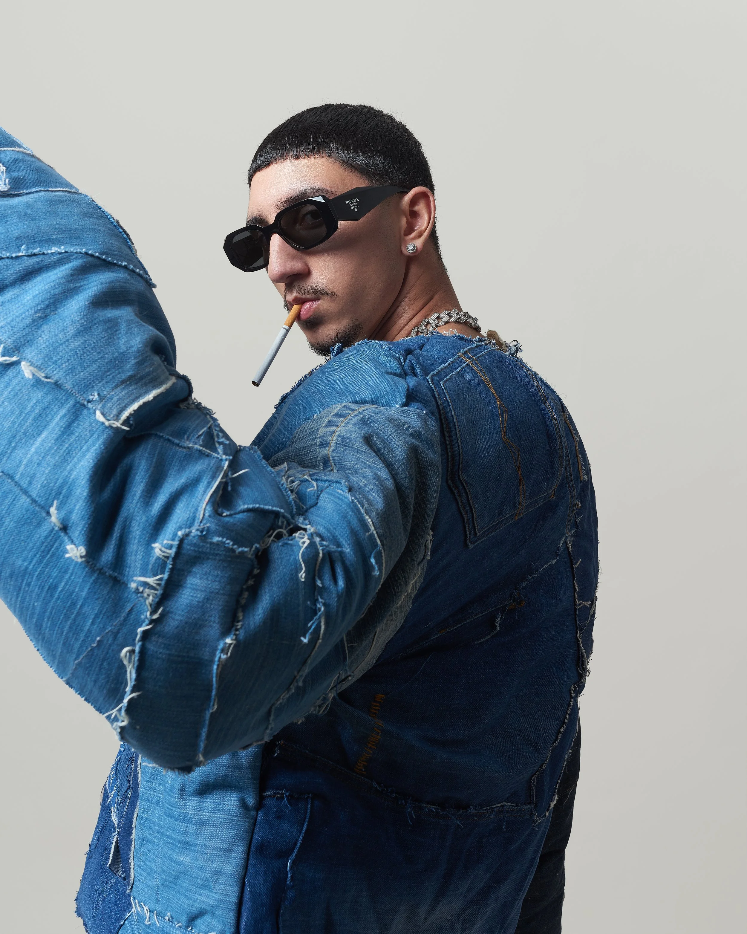 Jeune homme portant des vêtements en jeans déchirés, lunettes de soleil noires, un collier en chaîne, une boucle d'oreille en diamant, avec une cigarette dans la bouche, contre un fond blanc.