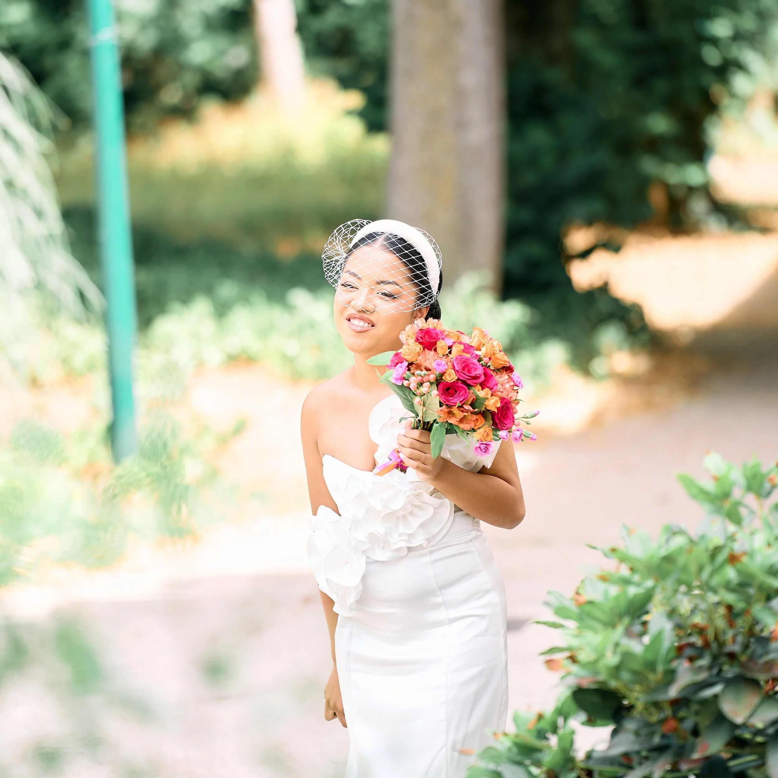 Jeune femme en robe de mariée blanche tenant un bouquet de fleurs colorées, portant un voile en filet et se tenant dans un parc en plein air.