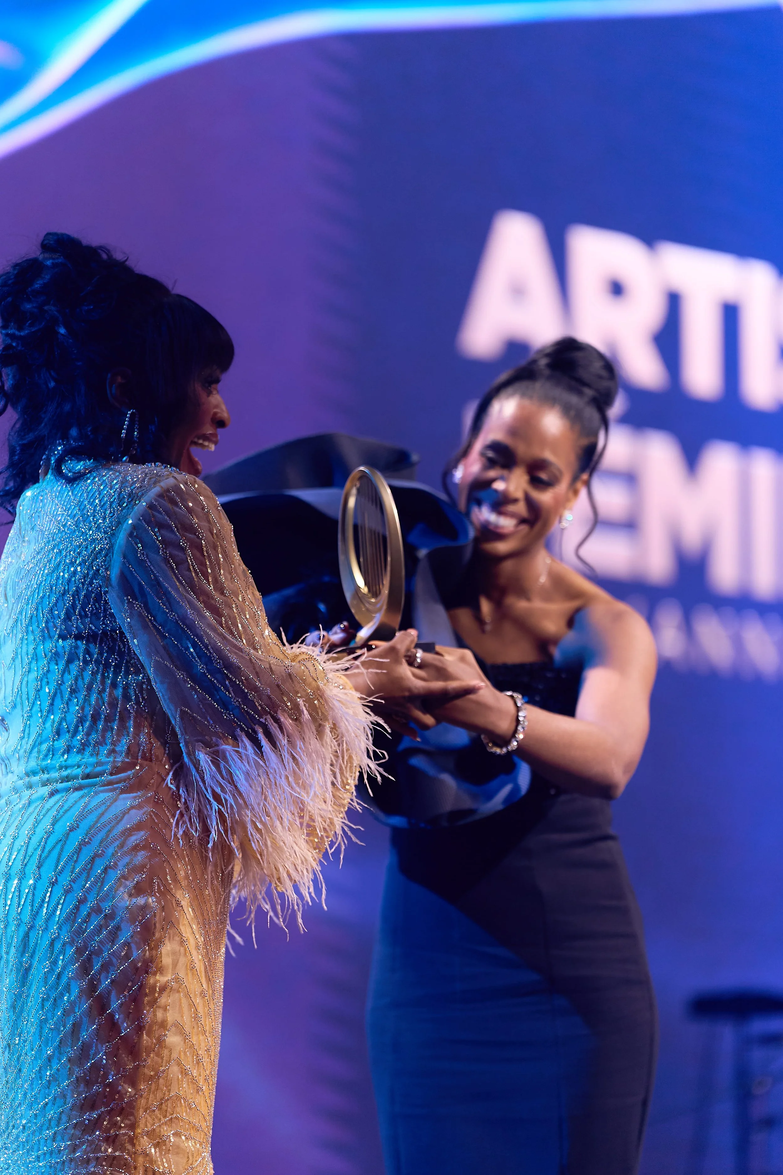 Deux femmes souriantes s'échangent un prix lors d'une cérémonie, en arrière-plan un panneau avec le texte 'Artiste Femme'.