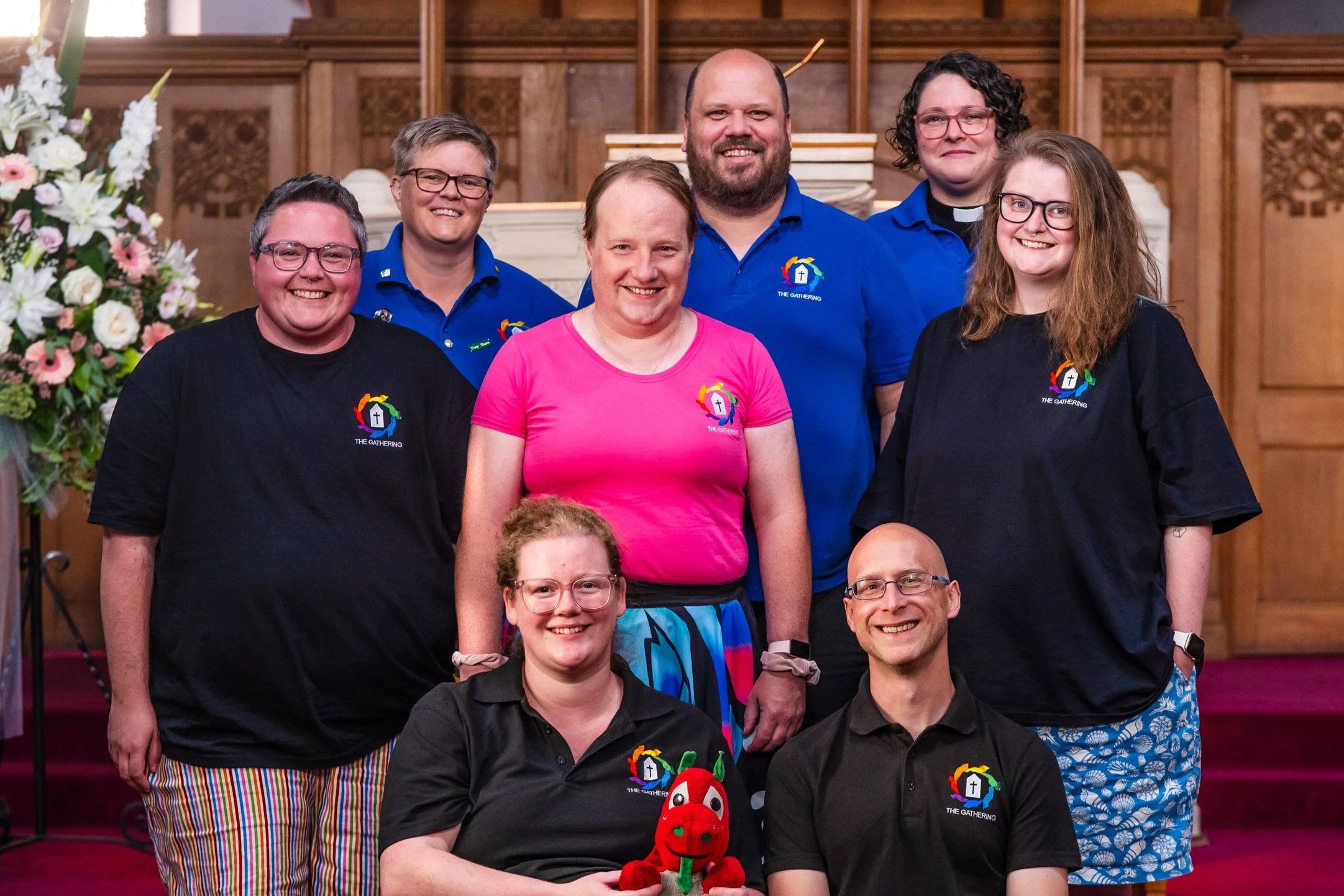 A picture of the trustees: four nonbinary people, two men, two women, and a small toy red dragon standing together and smiling