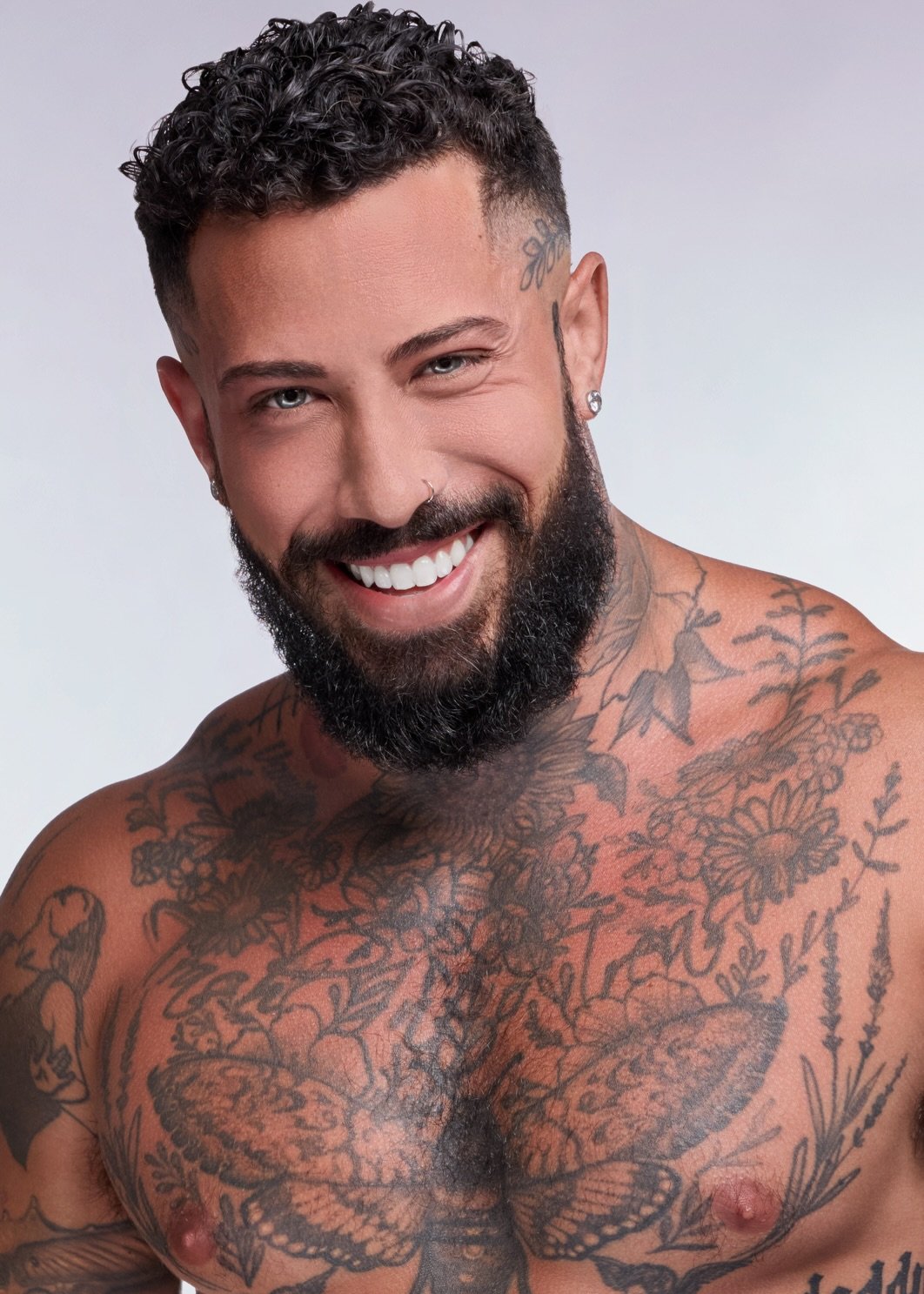 A smiling man with a beard and tattoos on his chest and face posing shirtless against a plain background.