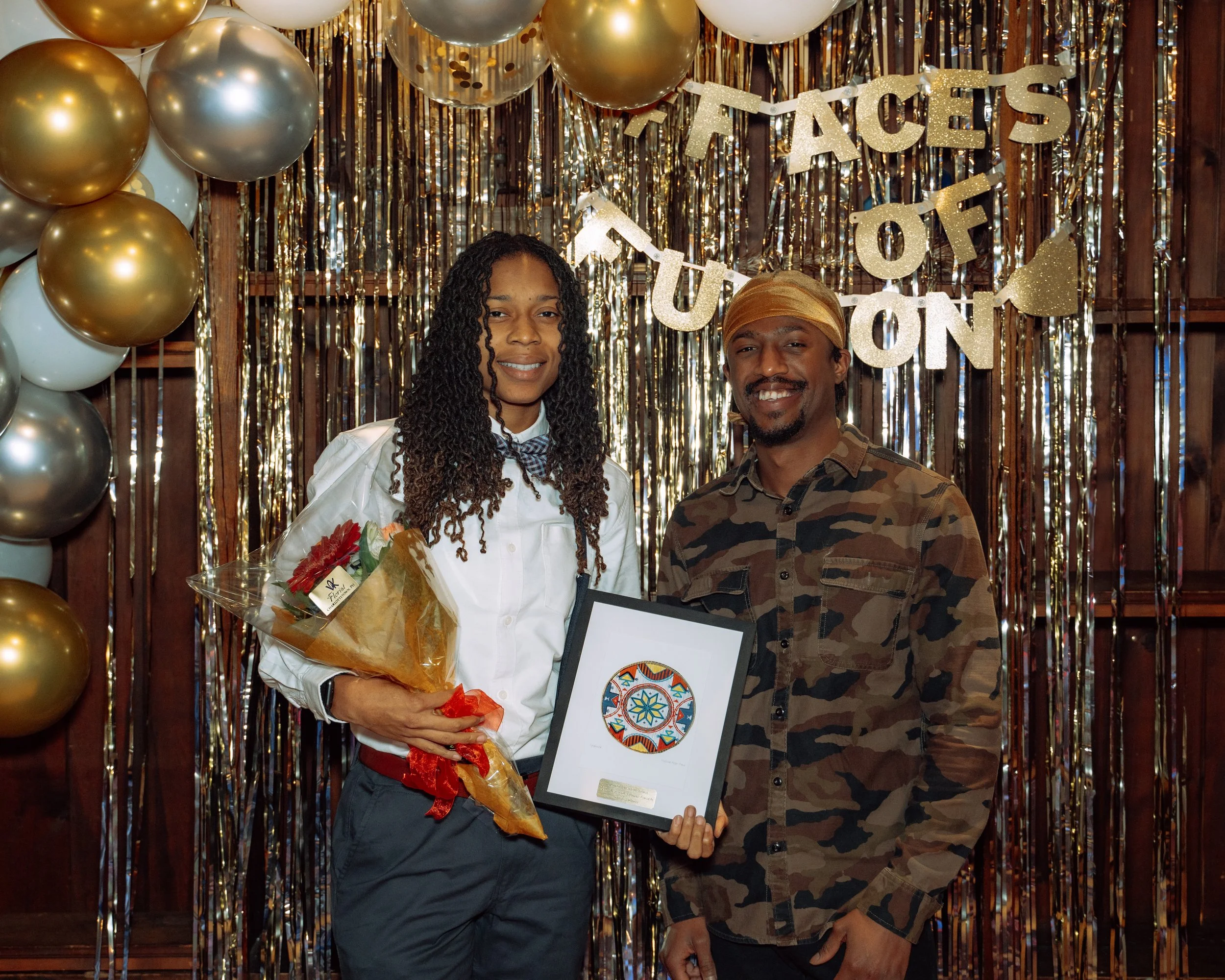 A woman and a man stand in front of a festive backdrop with balloons and metallic streamers. The woman holds a bouquet of flowers and a framed colorful artwork, while the man smiles nearby. The backdrop includes a sign that reads "Faces of Tutu".