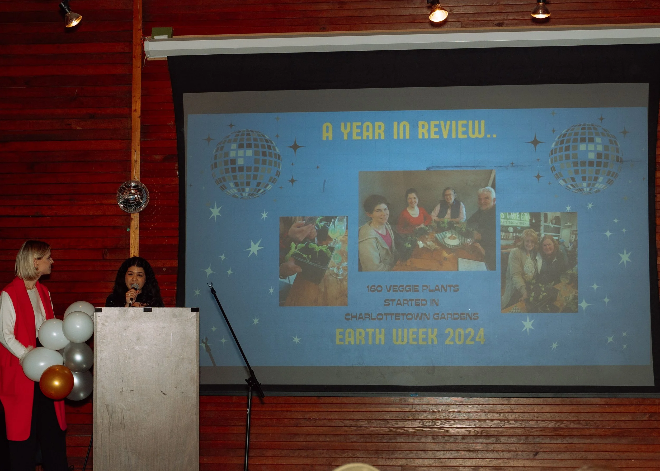 A presentation slide titled 'A Year in Review...' shows photos and notes about Earth Week 2024, including the start of a vegetable garden with 160 plants in Charlottetown Gardens, and features images of people, plants, and a disco ball decoration. Tw