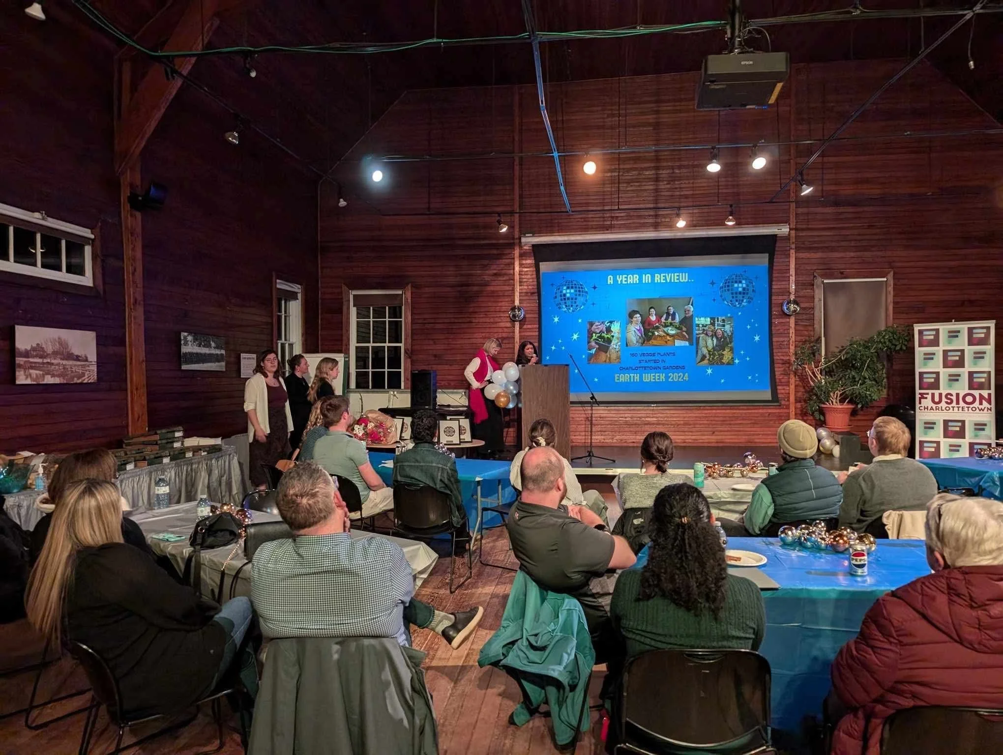 A group of people attending a presentation or event in a rustic wooden hall with a large screen displaying a slideshow about Earth Week 2024. There are tables decorated with balloons and snacks, and a person speaking at a podium.