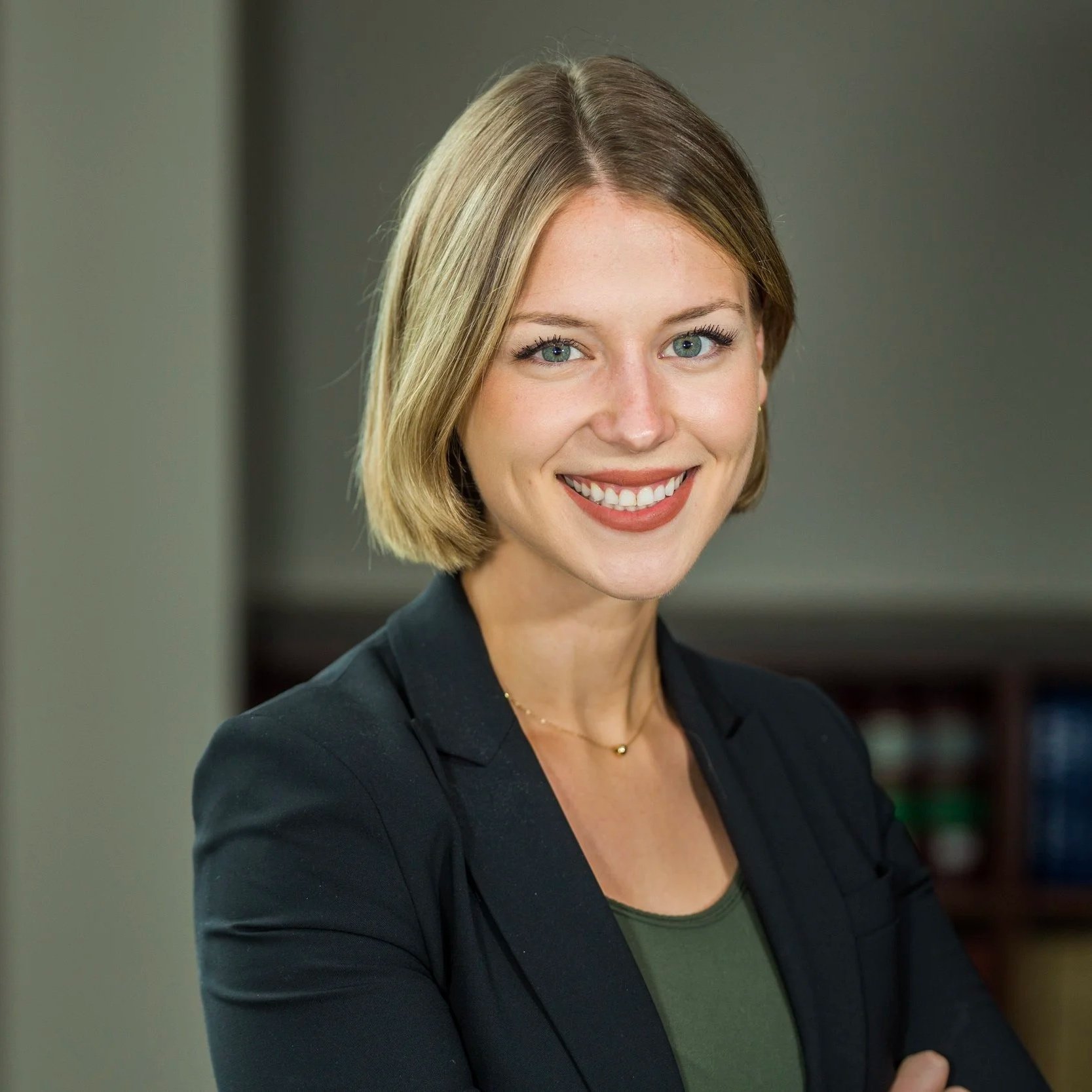 A smiling woman with short blonde hair, wearing a black blazer and a dark green top, standing indoors near a window.