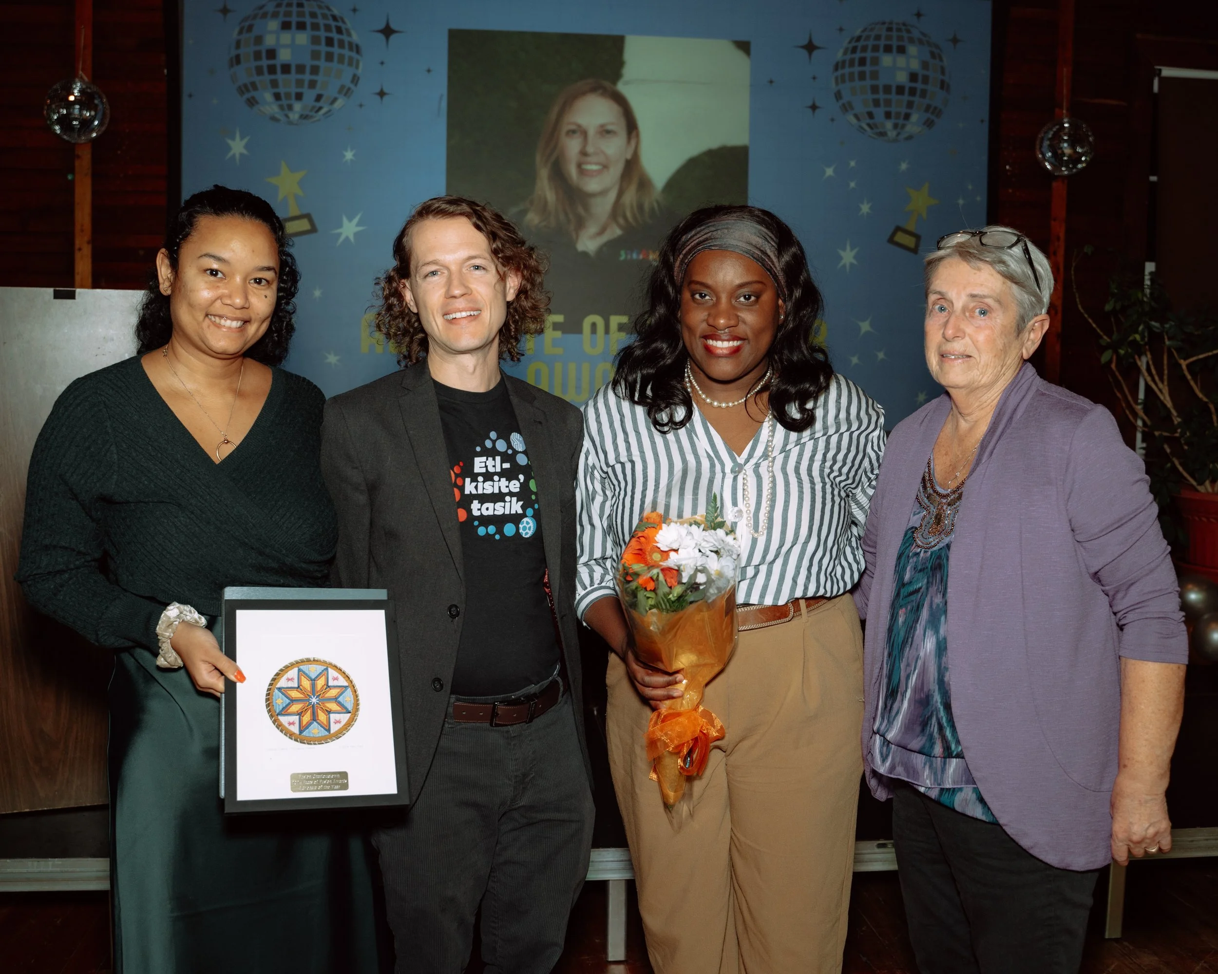 Four women standing together at an award ceremony, with one woman holding a framed award and a bouquet of flowers, smiling at the camera.