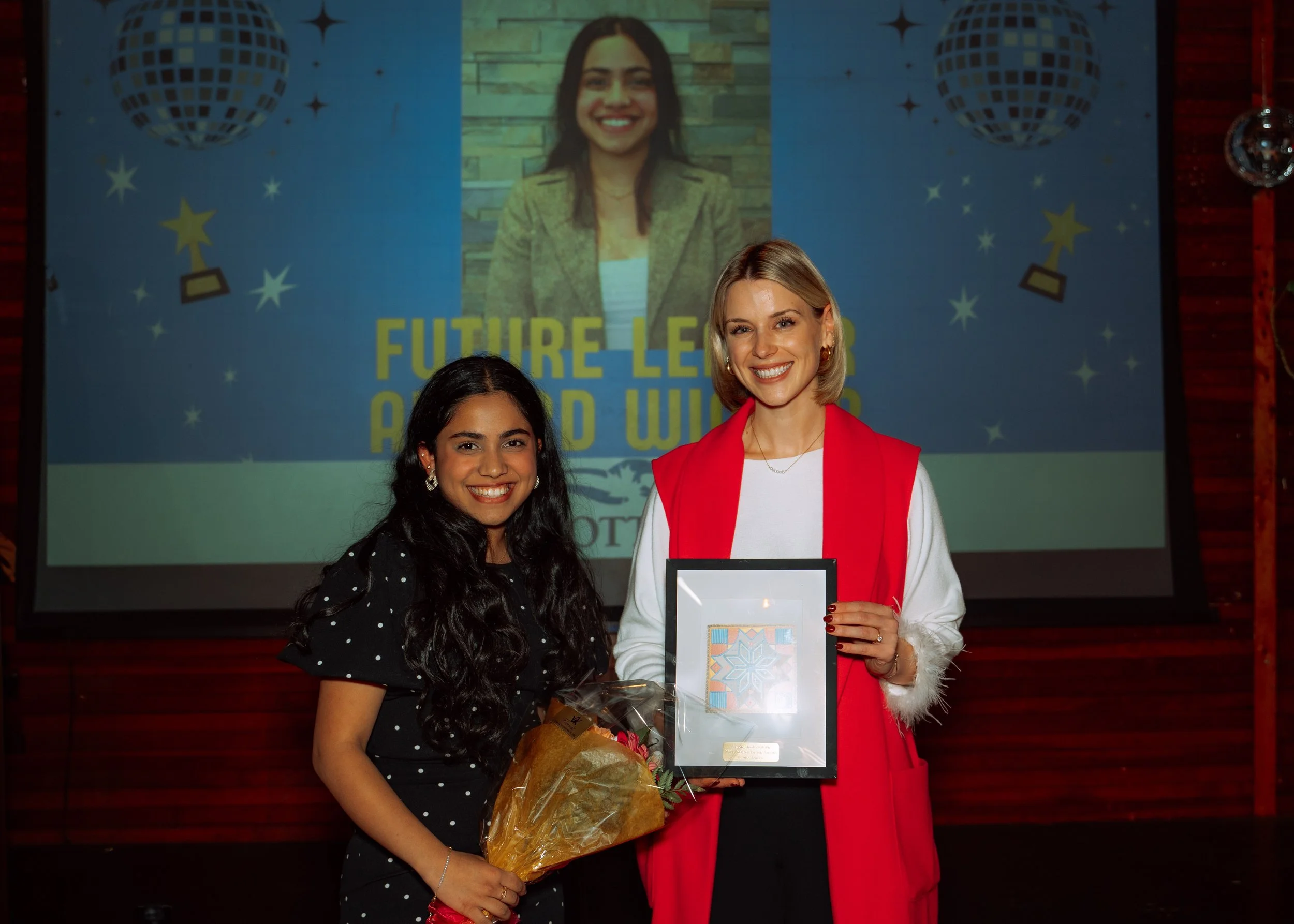 Two women standing on stage at an award event, one holding a framed certificate and the other holding a bouquet of flowers, smiling for the photo, with a large screen in the background displaying a woman's face and the words "Future Leaders Award Win