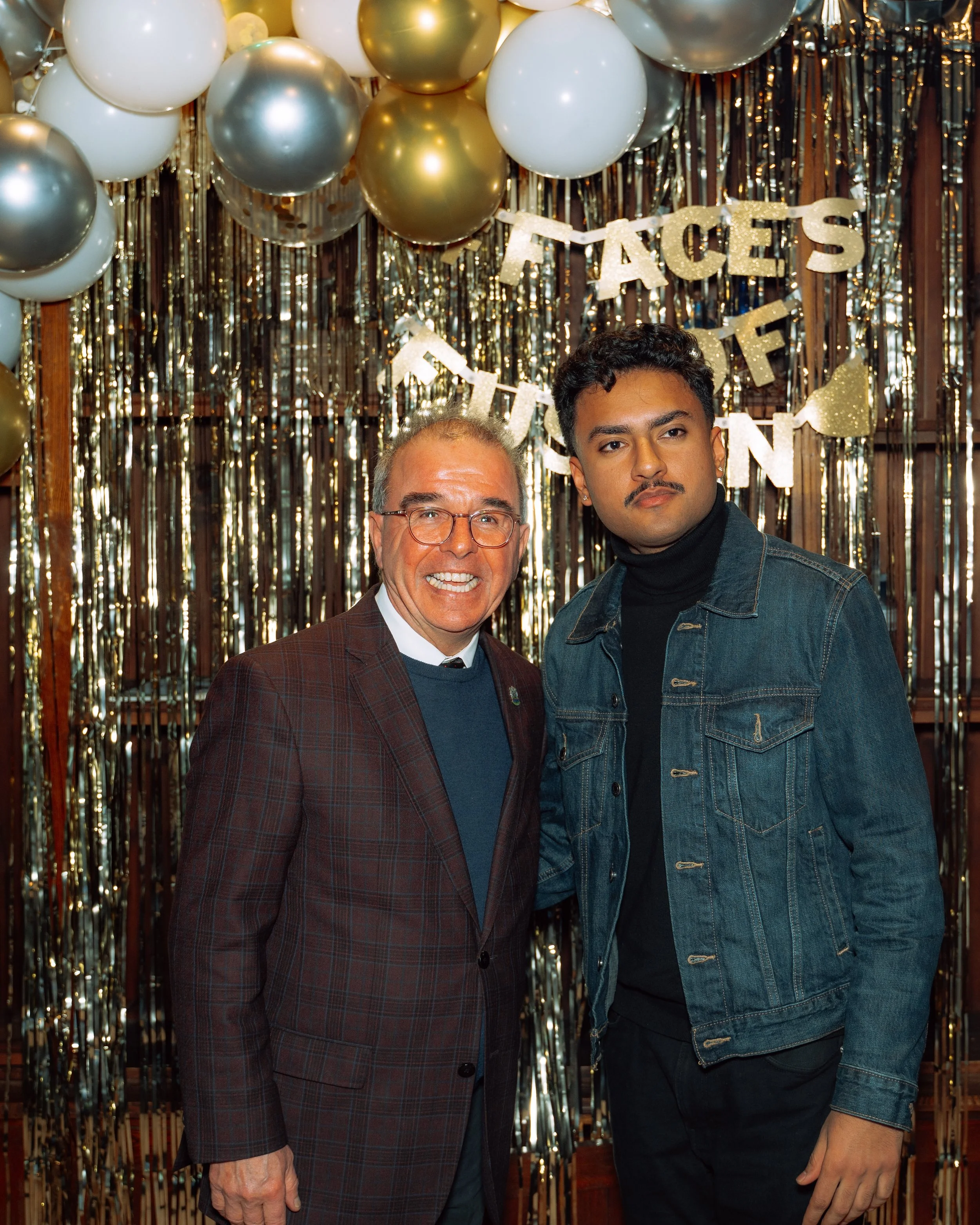 Two men standing together at a celebration with a gold and silver balloon backdrop and a banner that says 'Facets of'.