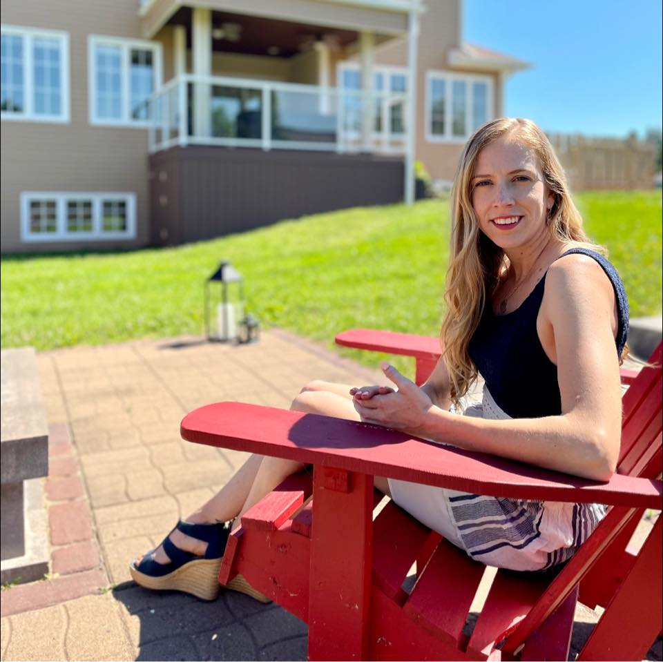 Young woman sitting on a red outdoor chair on a paved patio, smiling at the camera with a house and green lawn in the background.