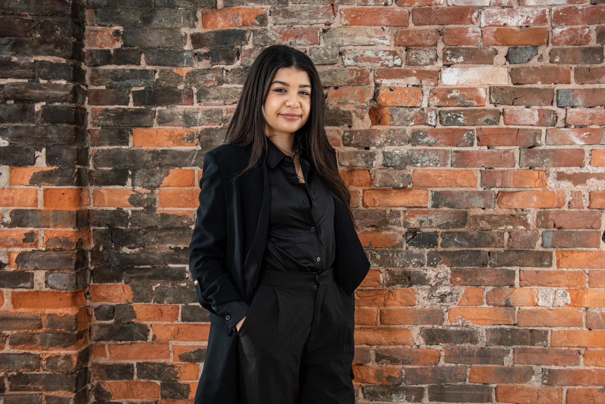 A woman with long dark hair wearing a black blazer and black pants, standing with her hands in her pockets against a brick wall.