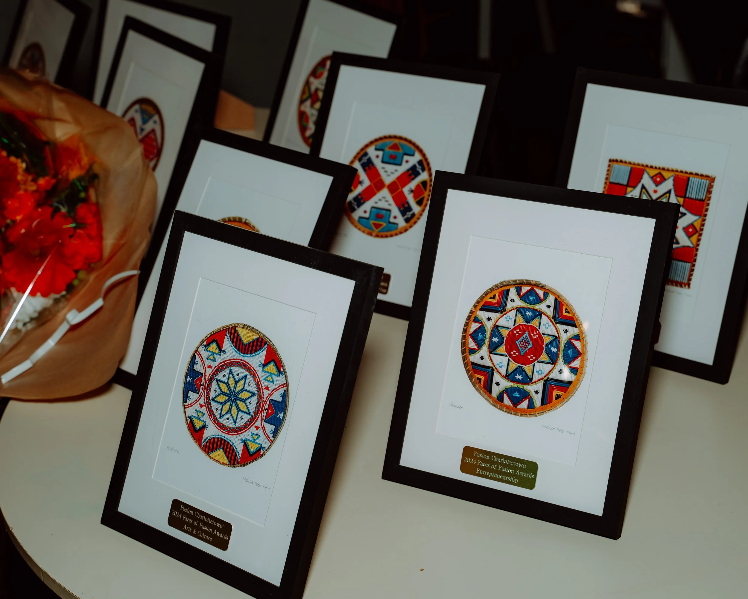 Several framed pieces of artwork with colorful geometric and circular patterns displayed on a table. A bouquet of flowers is partially visible on the left side.