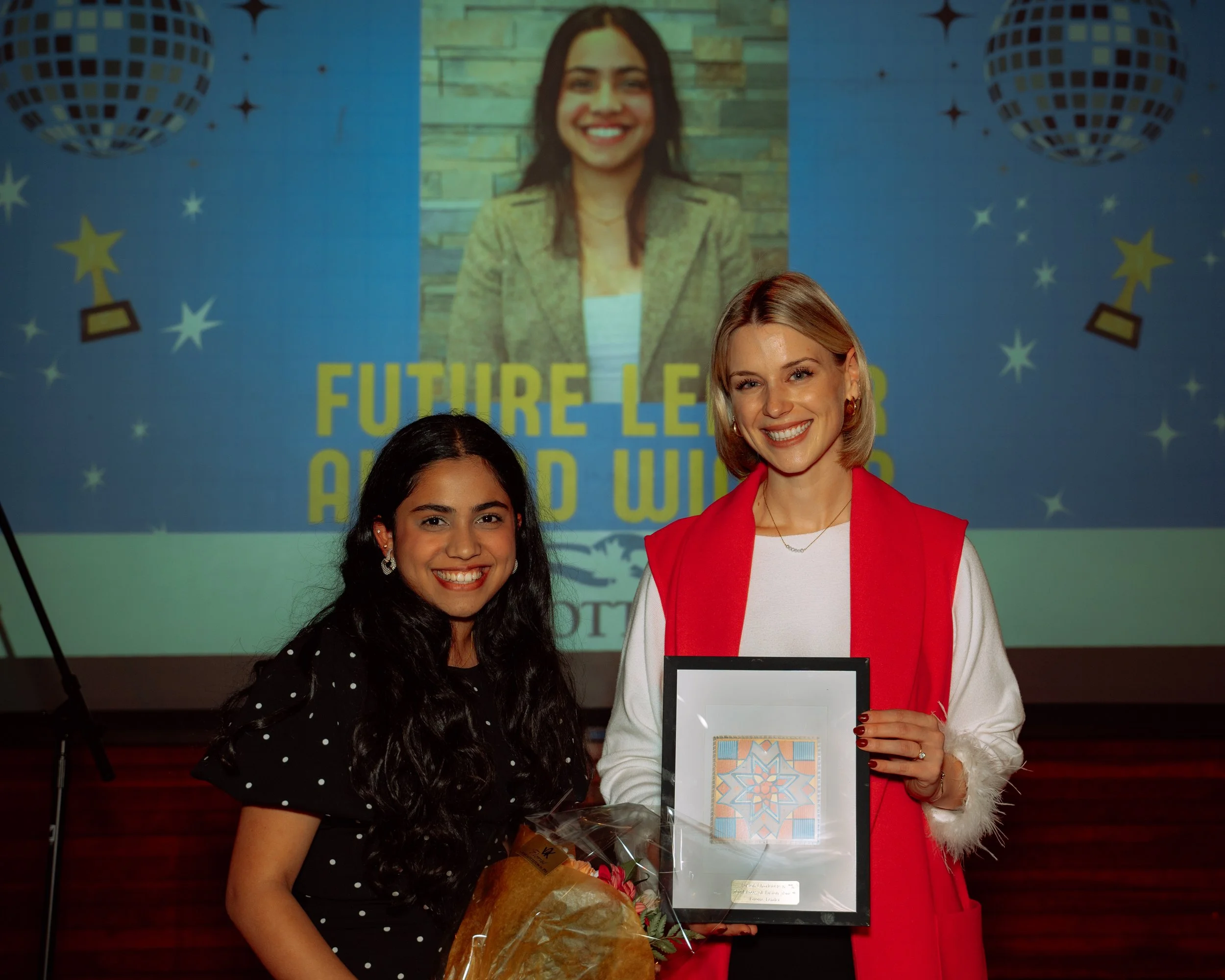 Two women standing together, smiling at the camera. The woman on the right, with blonde hair, is wearing a red vest over a white top and holding a framed artwork. The woman on the left, with long dark hair, is dressed in a black polka dot dress and h