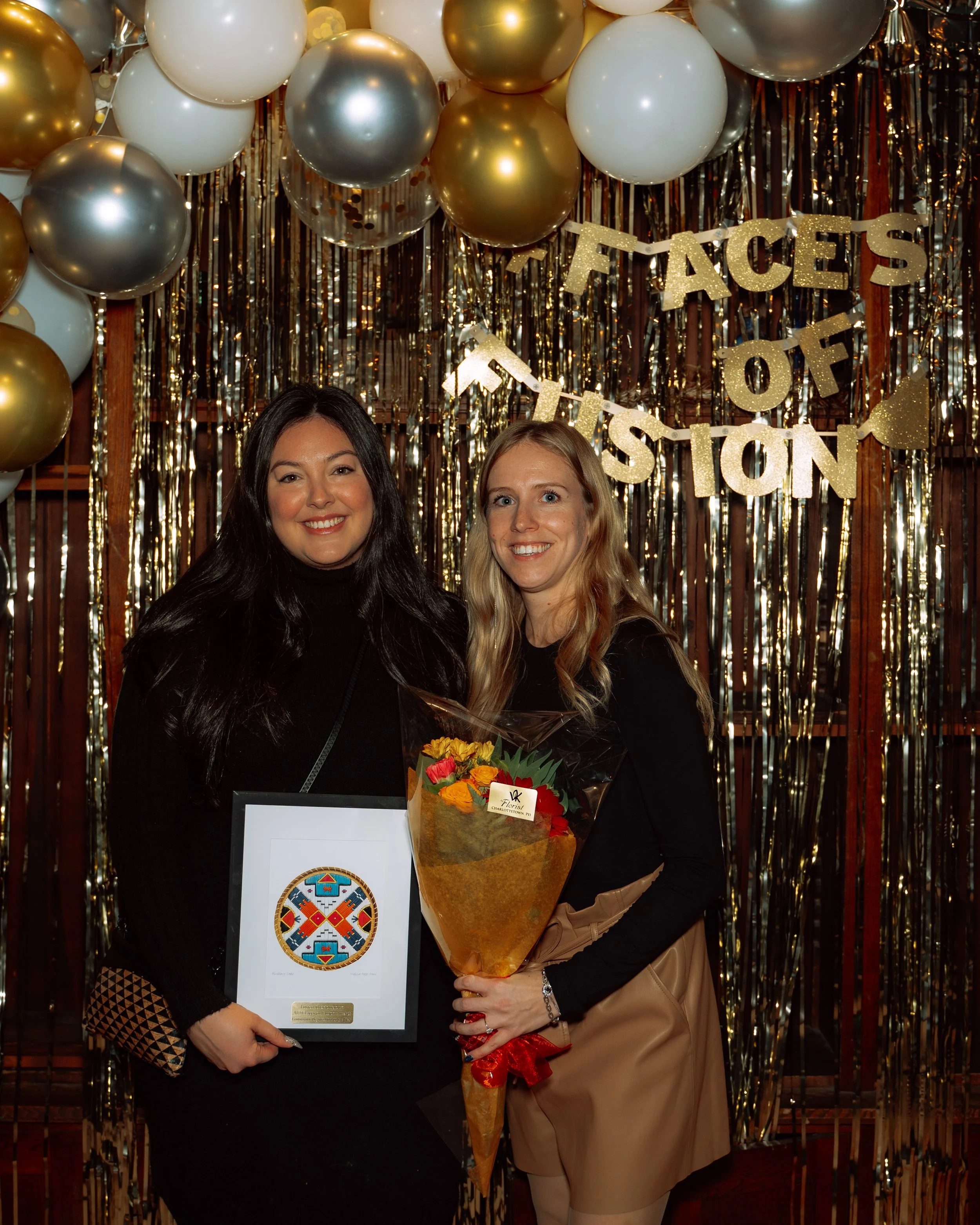 Two women standing in front of a gold and white balloon backdrop with a 'Faces of The Station' banner. One woman is holding a framed certificate or artwork, the other a bouquet of flowers. Both are smiling and dressed in black.