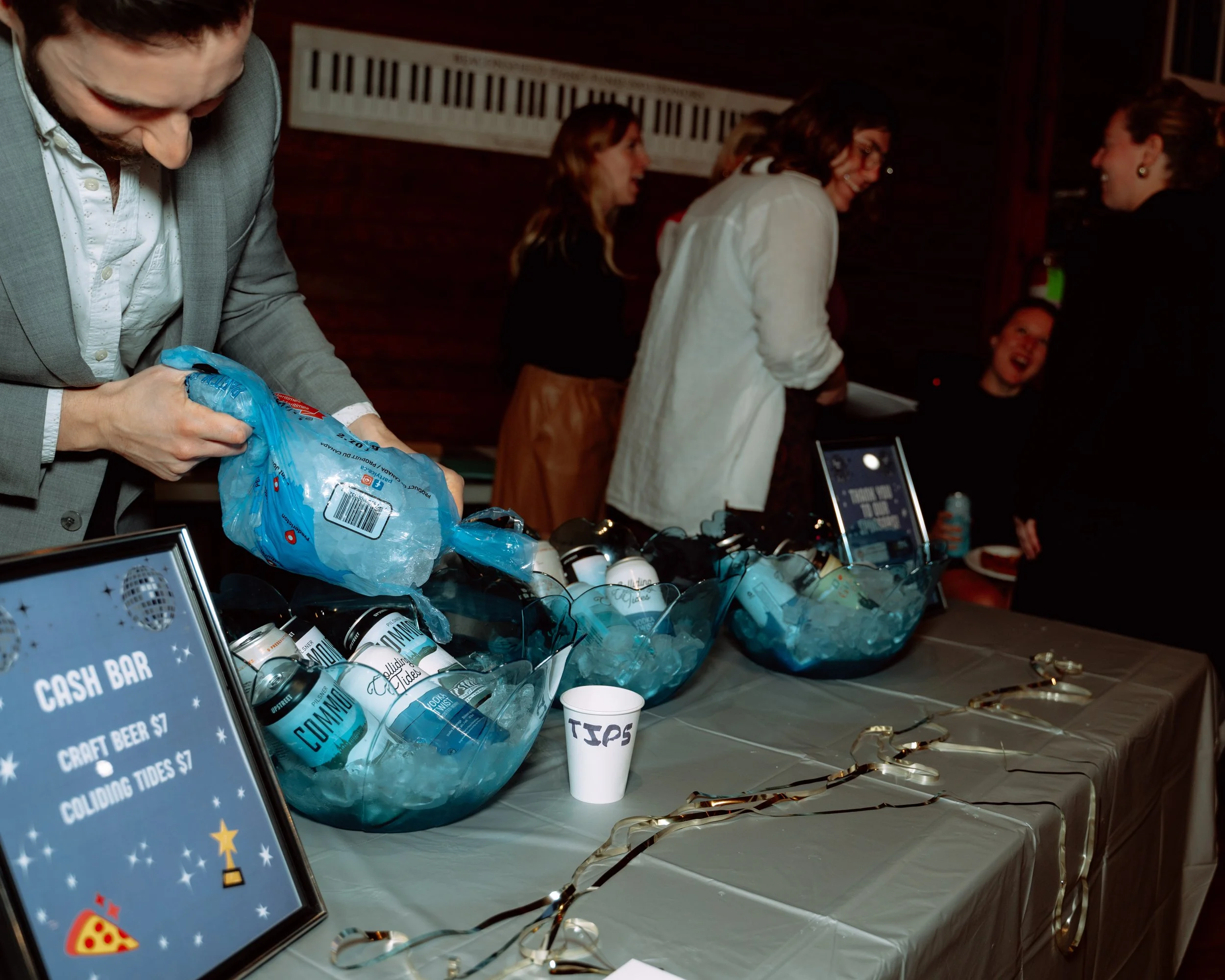 A man in a gray suit filling a plastic bag with bottled drinks at a party table decorated with gold streamers. In the background, people are laughing and talking, and a sign lists craft beer and colding tides for sale.